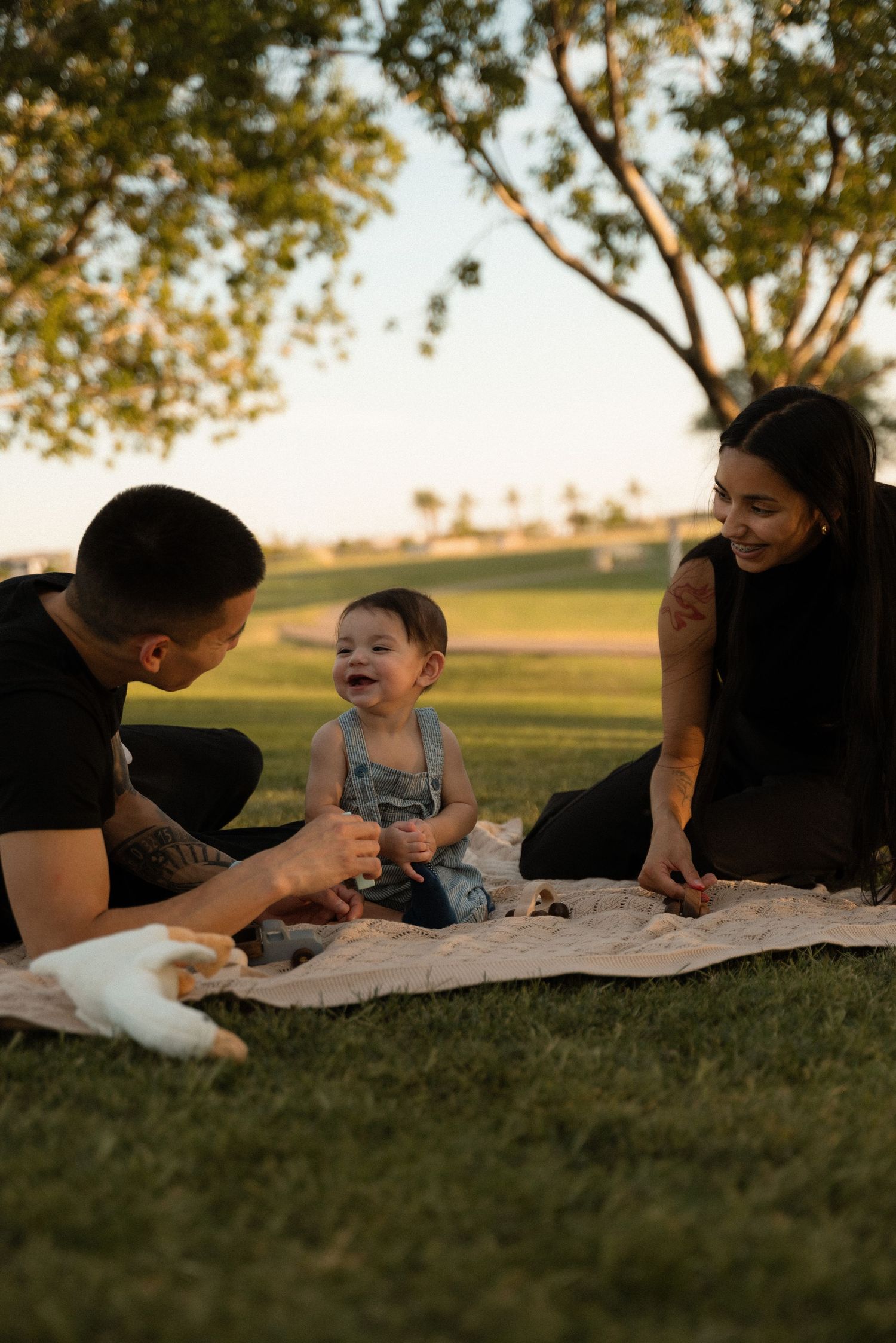 A happy family enjoying a picnic on a blanket in a park at sunset with their laughing baby.