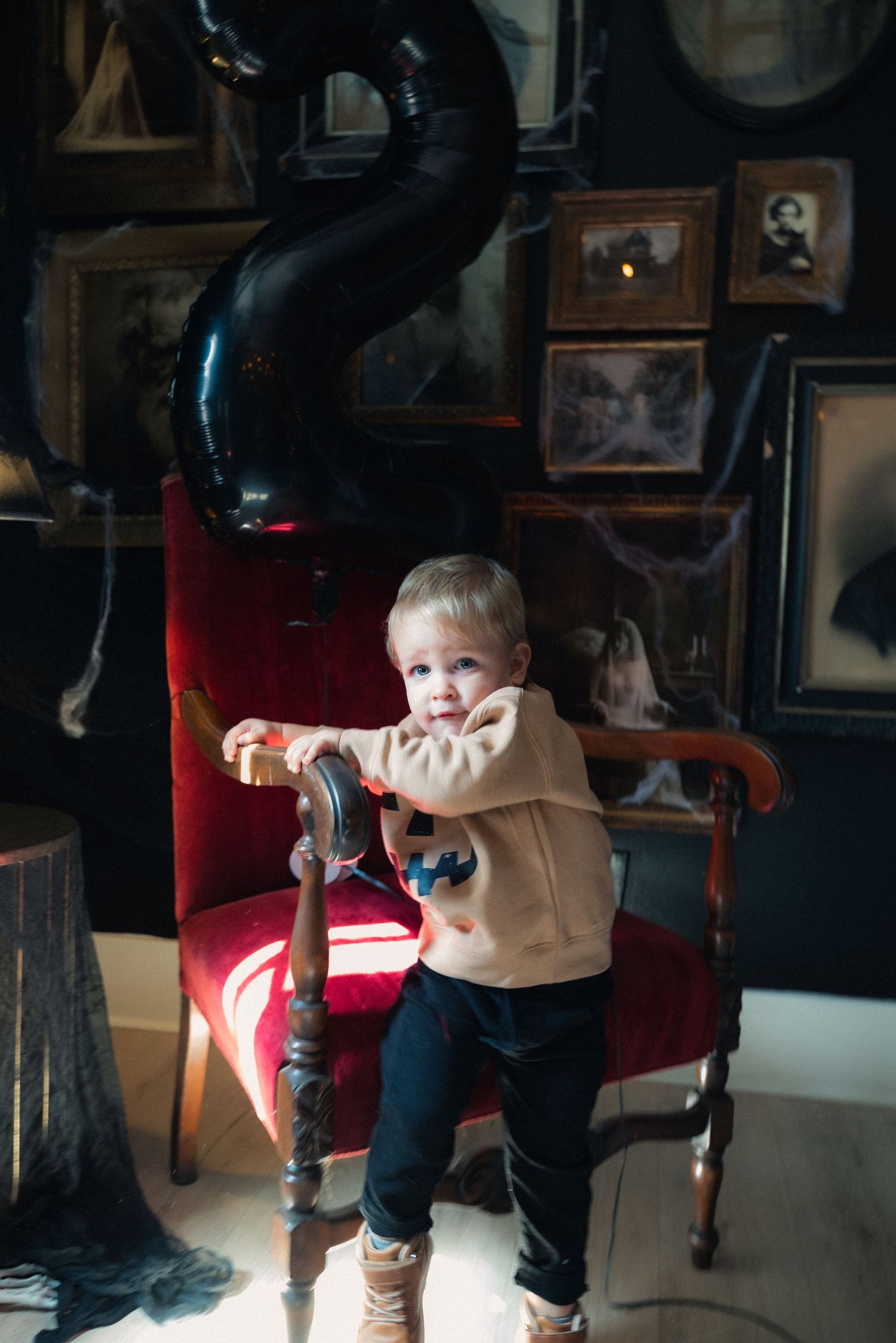 A young child stands on a red vintage chair in a dimly lit room filled with framed photos and antique decor.