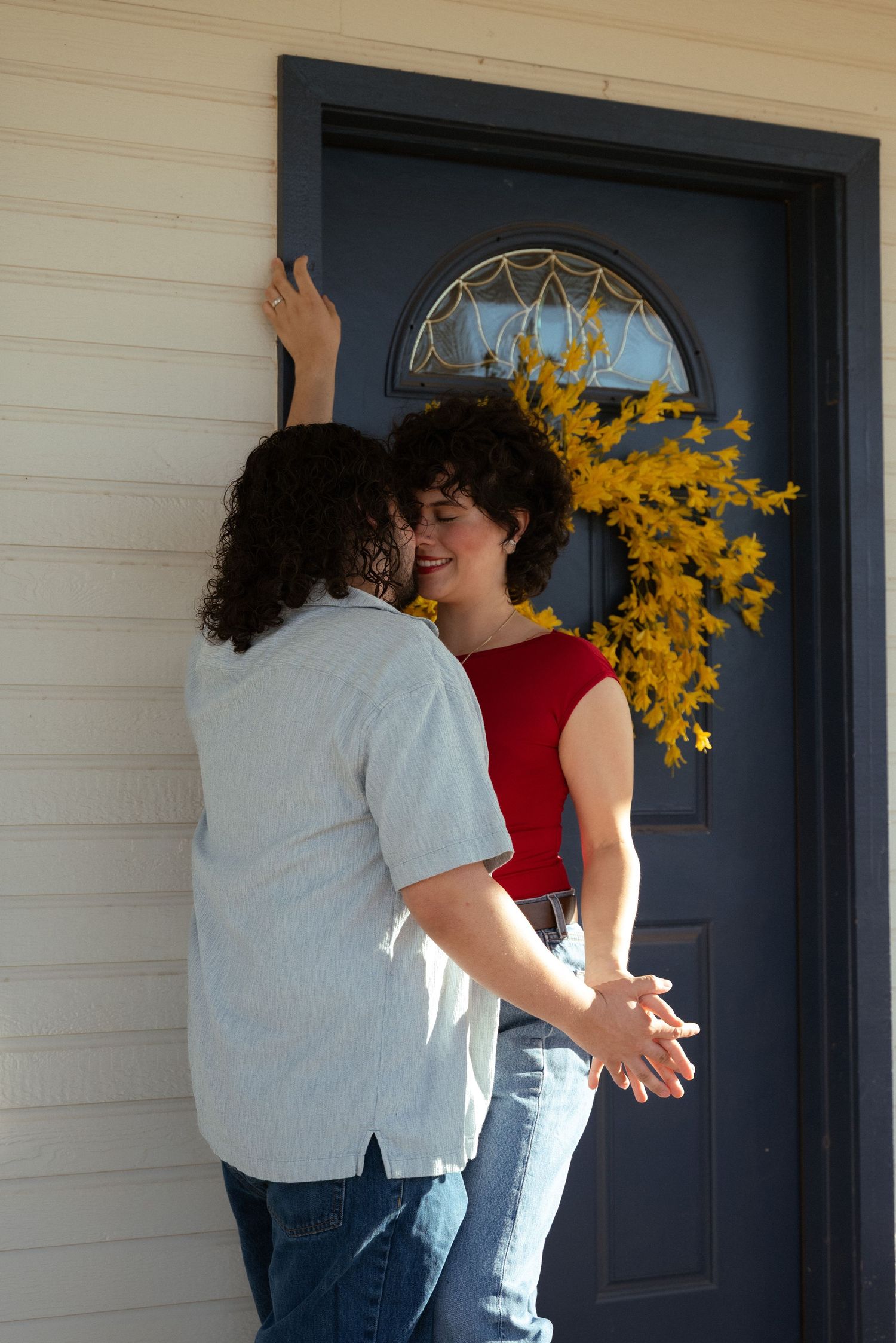 Couple sharing a romantic moment at a navy blue front door decorated with a yellow floral wreath on a sunny day.