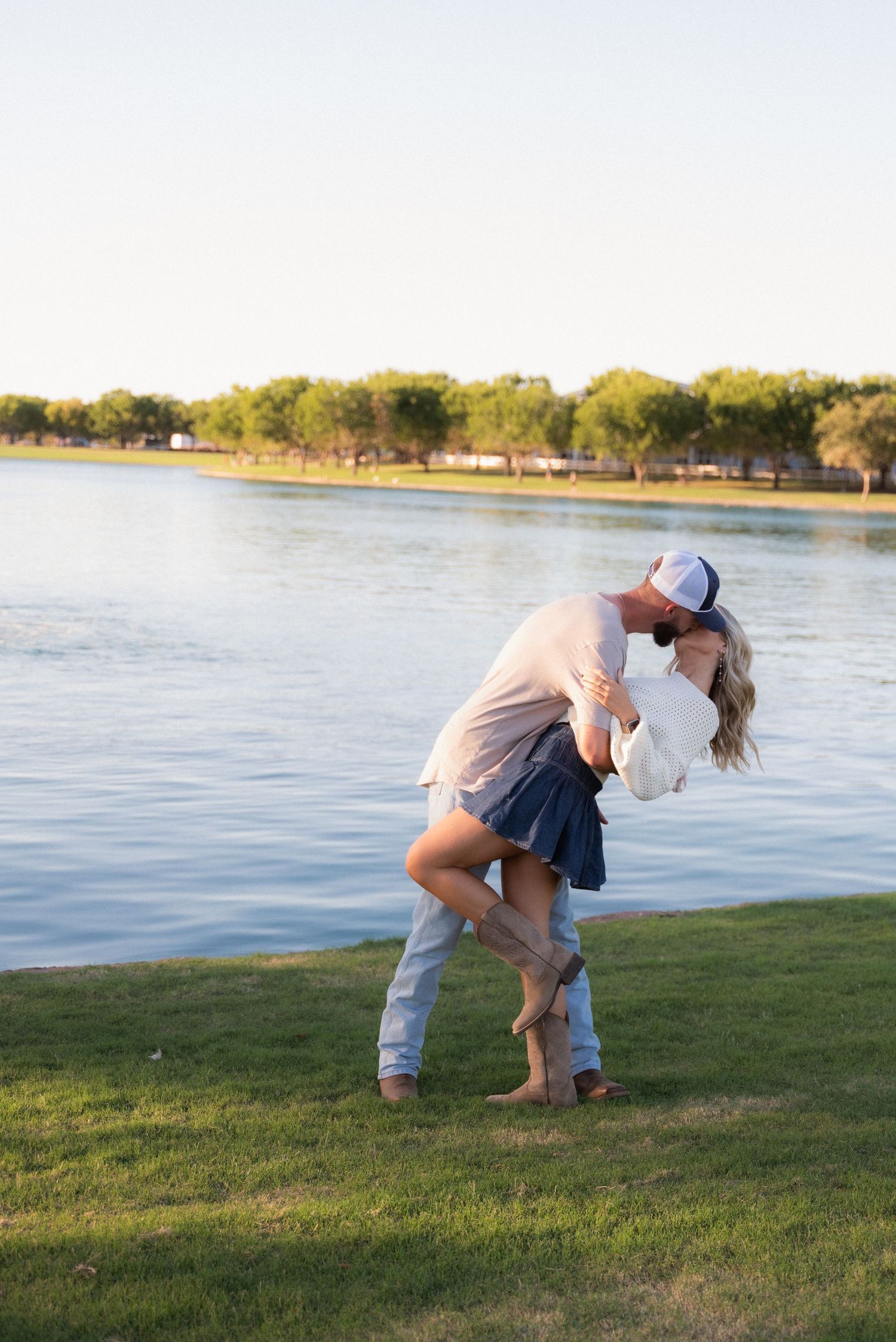 Couple embraces in romantic pose by lakeside at sunset on grassy shore.