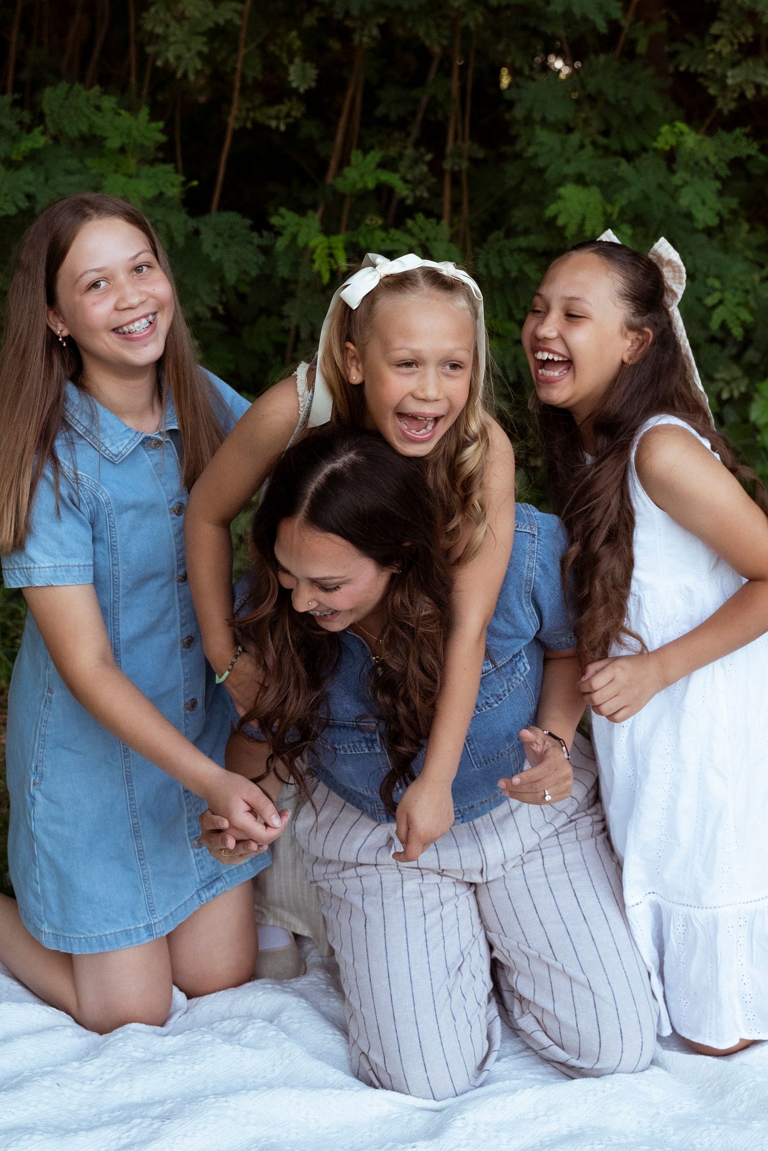 Four friends in casual dresses share a joyful moment laughing together while posing on a white blanket outdoors.