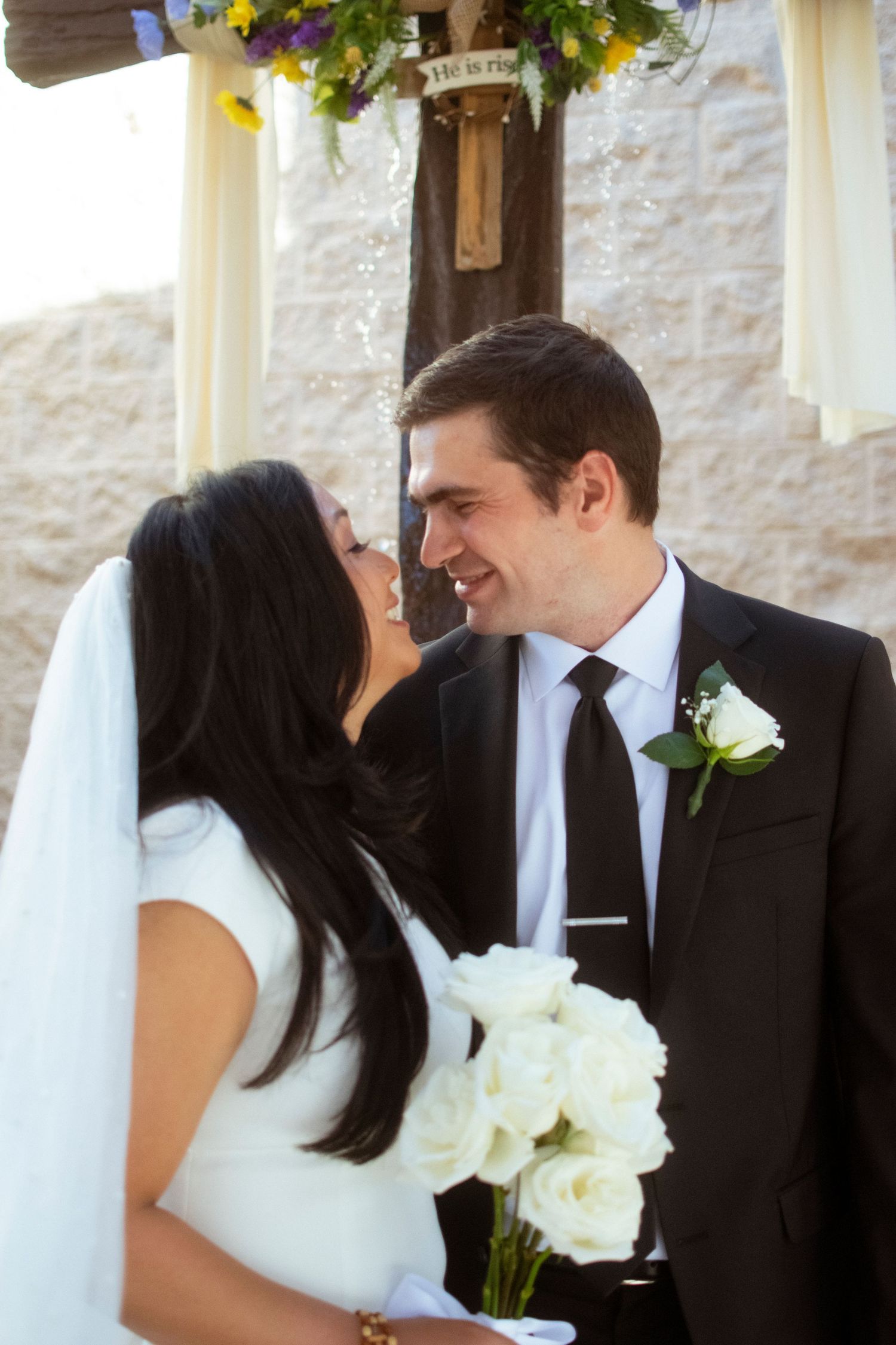 A bride in white holds a bouquet of white roses while sharing an intimate moment with her groom at their wedding.