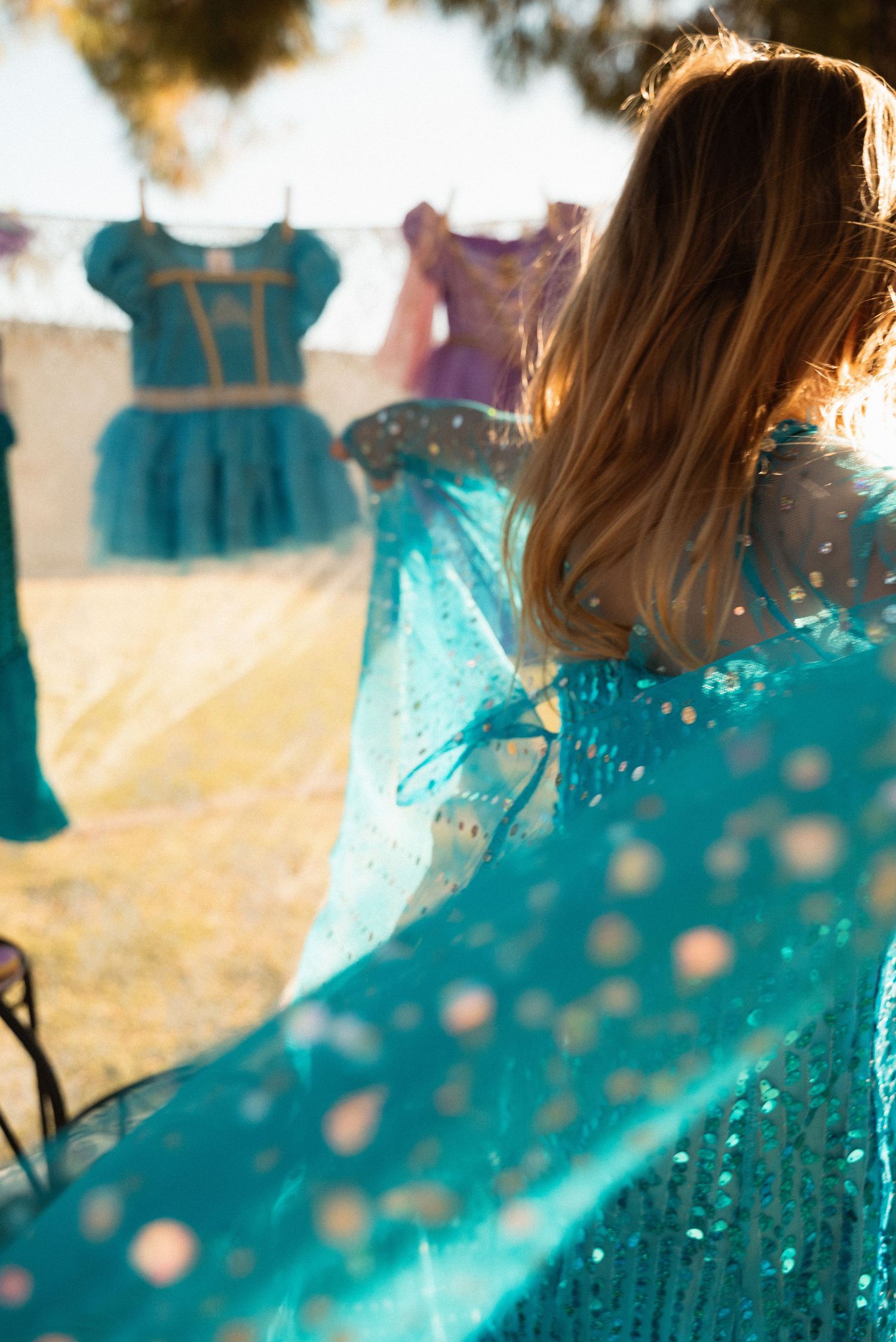 Girl in teal sequin dress holds out skirt outdoors, with colorful princess dresses hanging on a clothesline behind her.