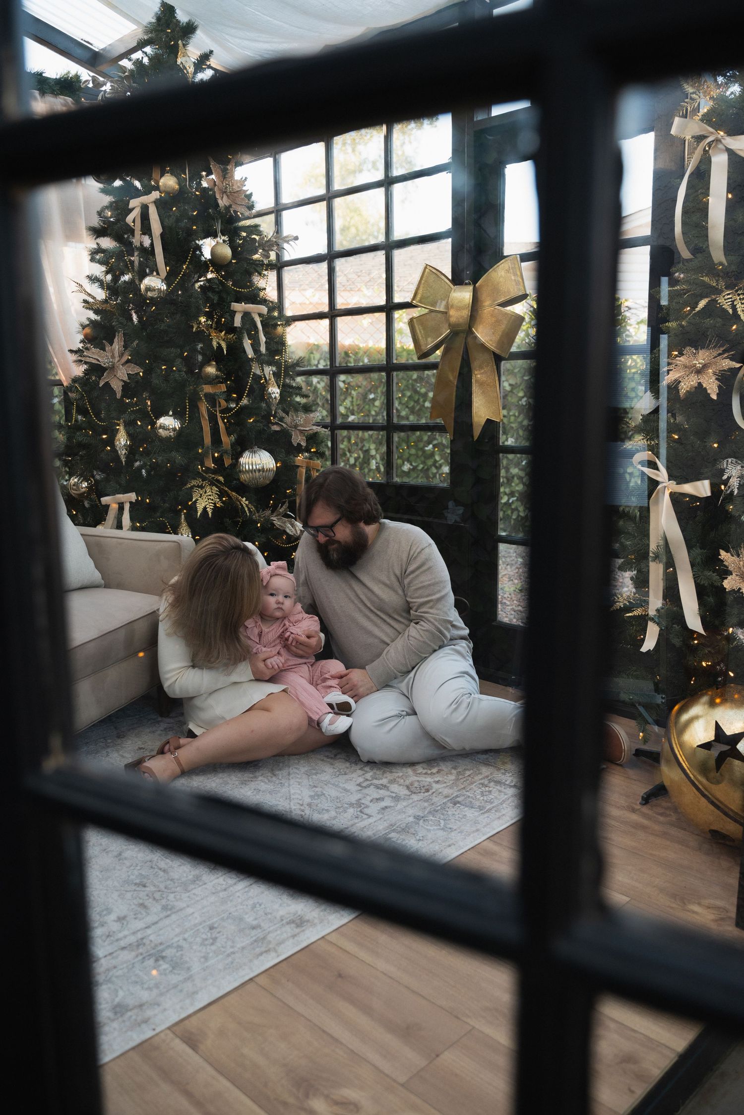 Family with baby sitting on floor by decorated Christmas trees, viewed through window with gold bow.