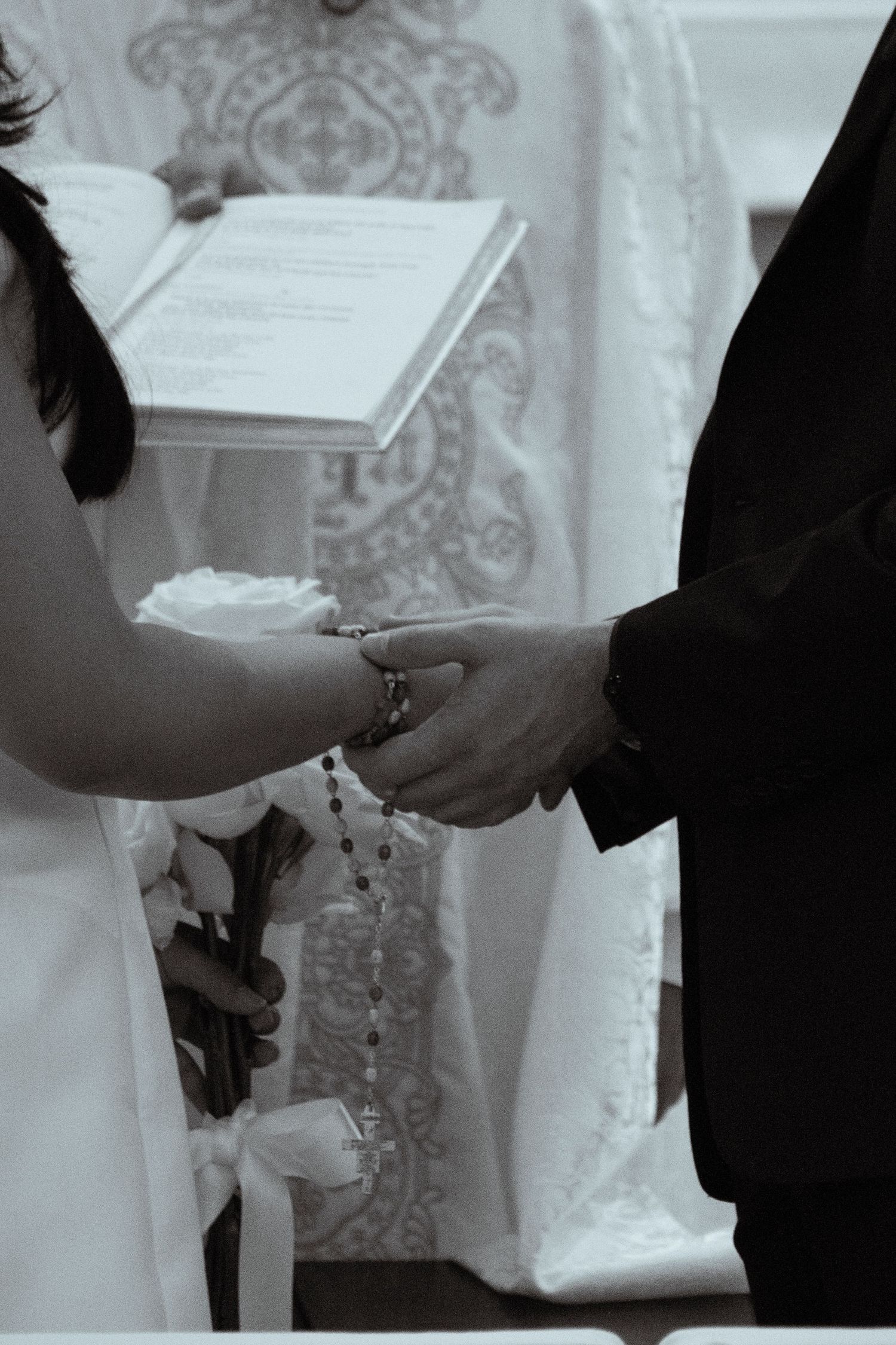 A black and white image of hands clasped together during a wedding ceremony with an open book in the background.