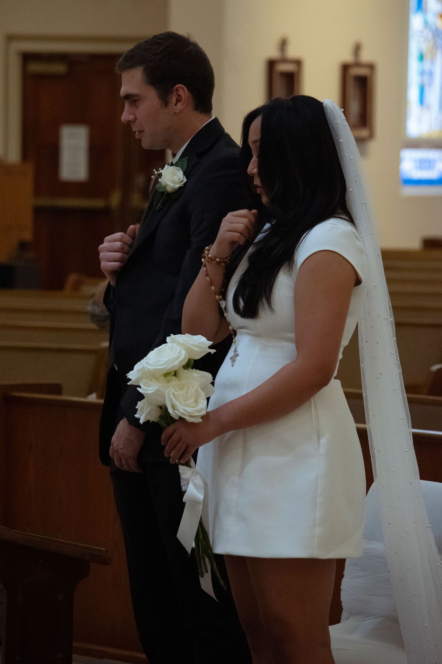A couple stands together during their wedding ceremony in a traditional church setting with wooden pews and stained glass.
