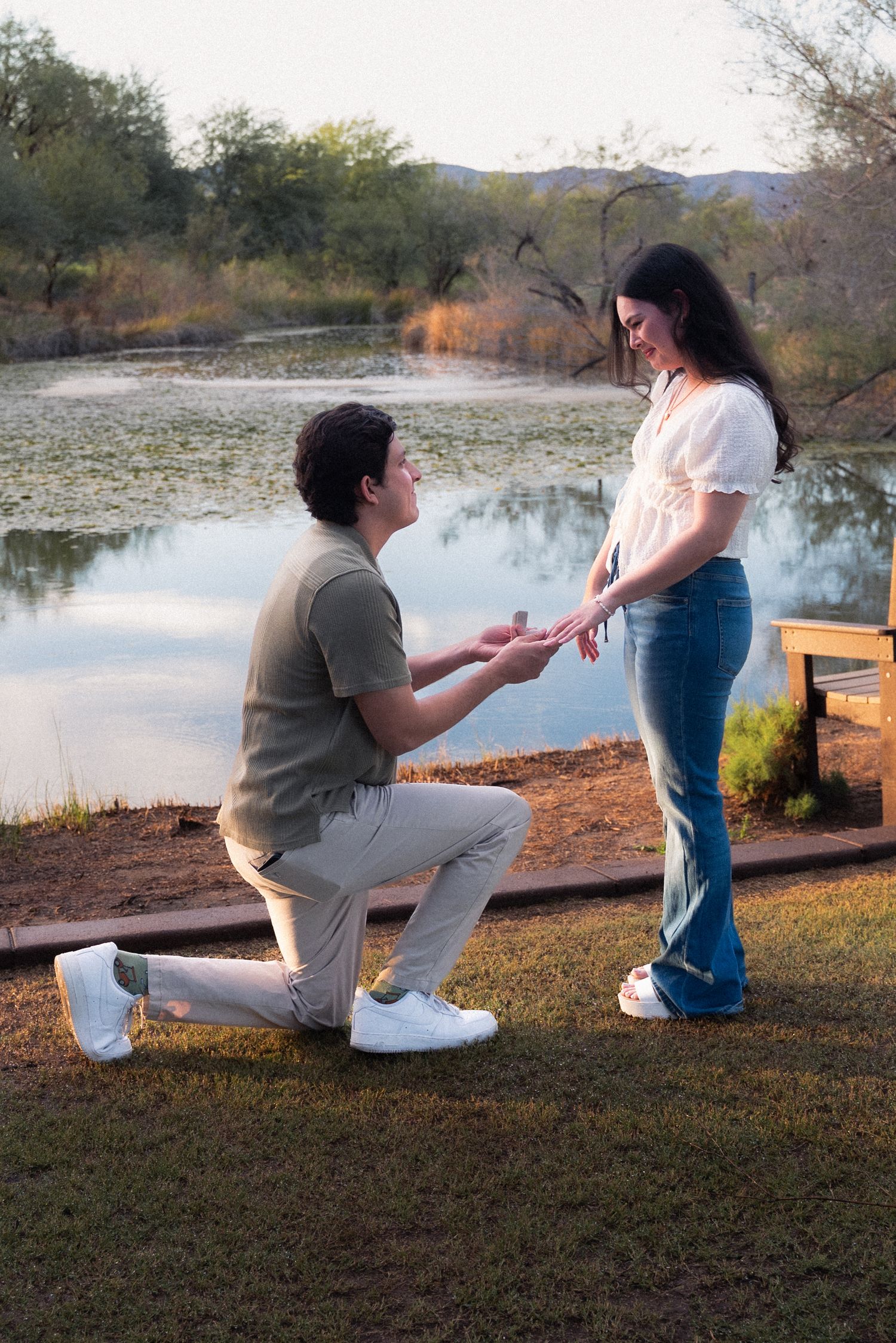 A romantic proposal scene unfolds by a serene lake during golden hour as a couple shares a special moment on the grass.