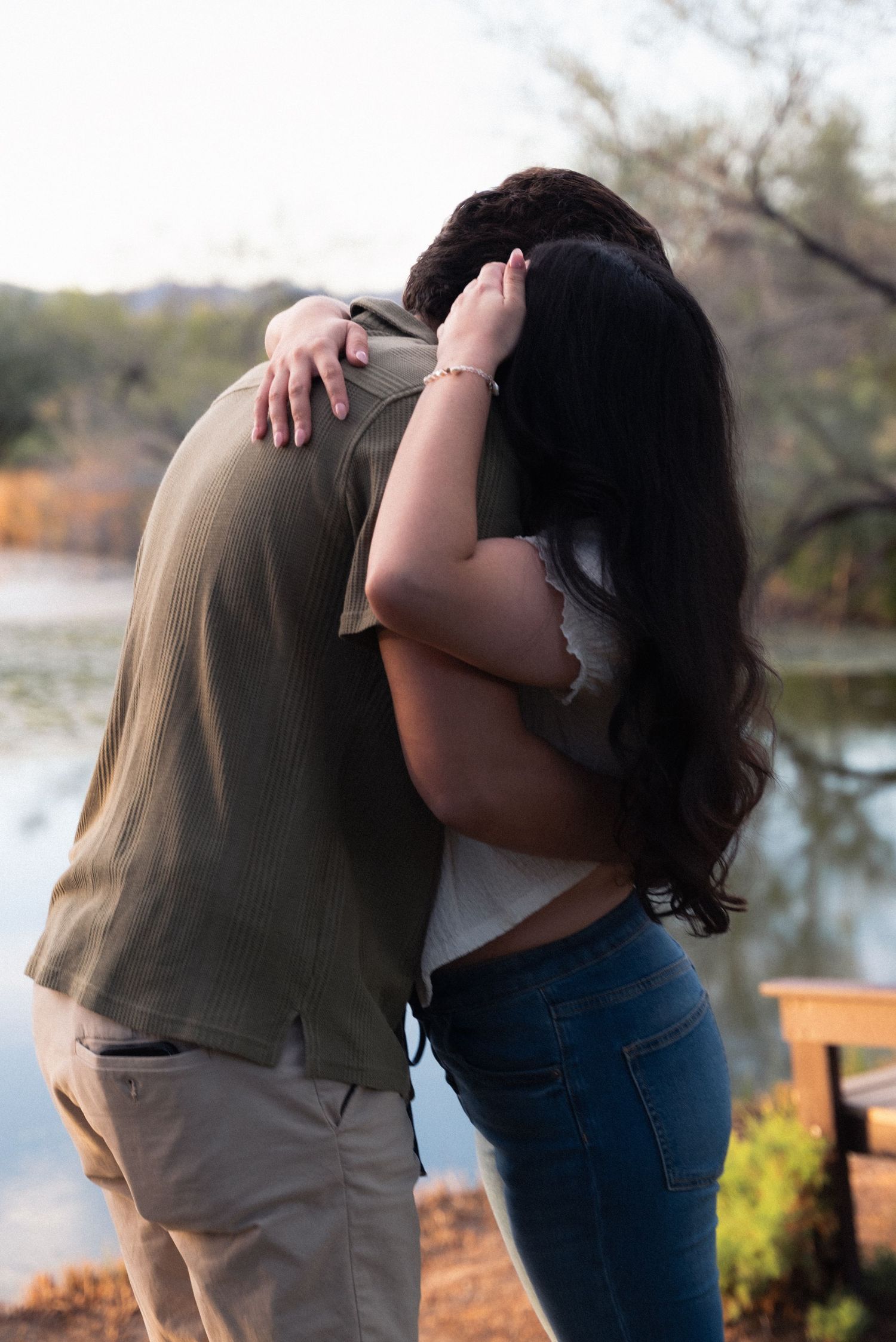 A couple embraces outdoors near a lake during autumn with fall foliage visible in the background.
