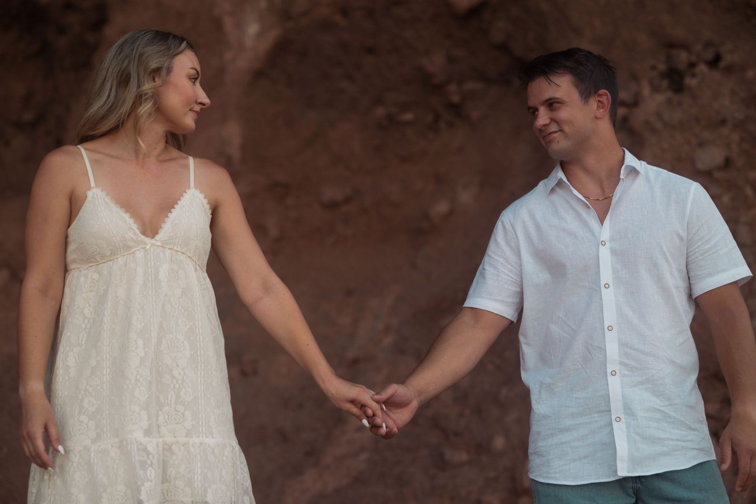 Couple in white attire holding hands against a red rock backdrop during an outdoor engagement photo session.