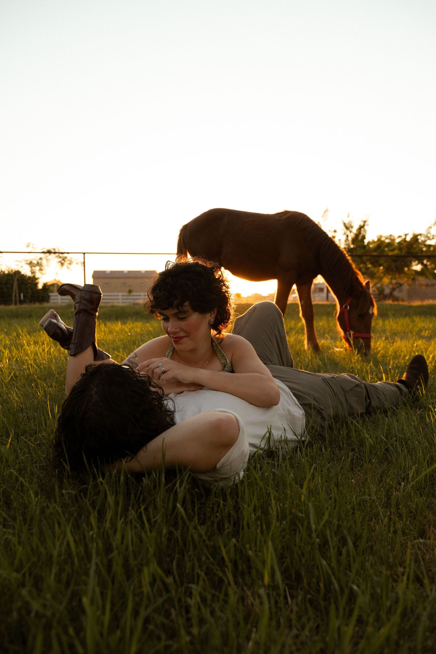 Couple lying in green grass at sunset with a brown horse grazing peacefully in the background.