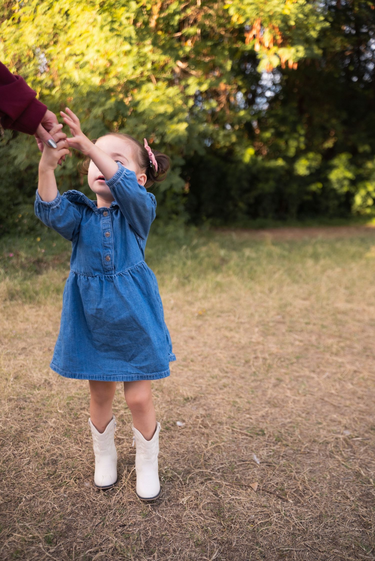 A small child in a denim dress and white socks stands outdoors in autumn with yellow foliage in the background.