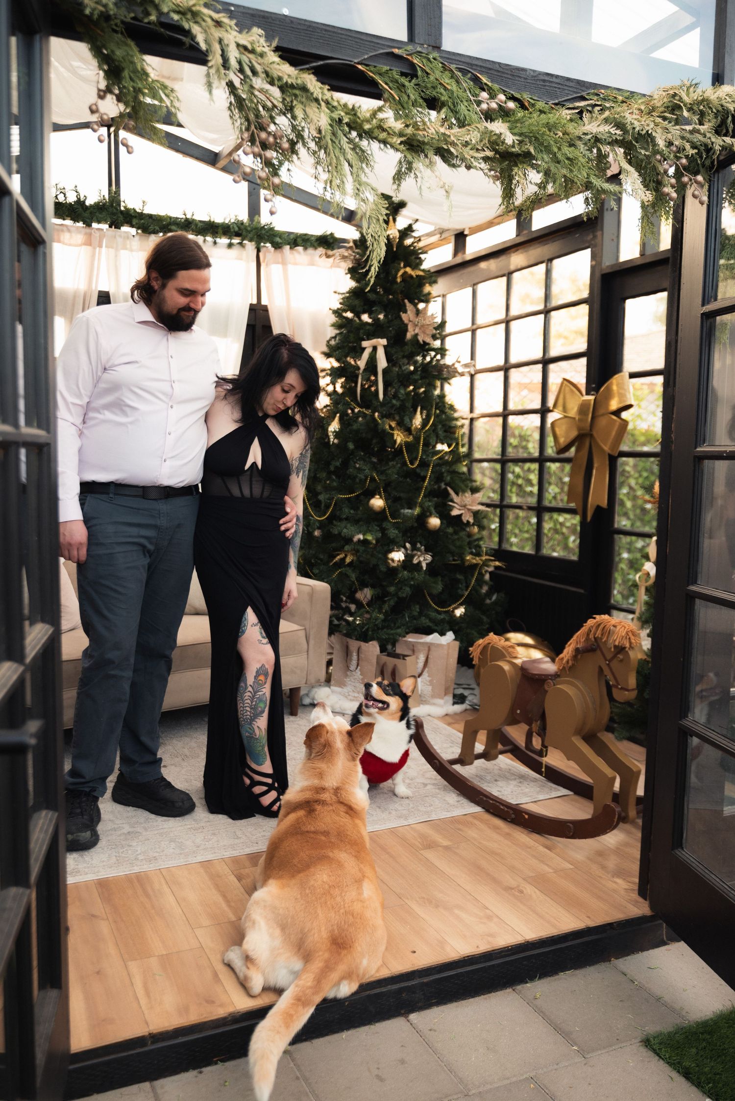 Couple in elegant attire stands by decorated Christmas tree with golden retriever and rocking horse in festive glass room.