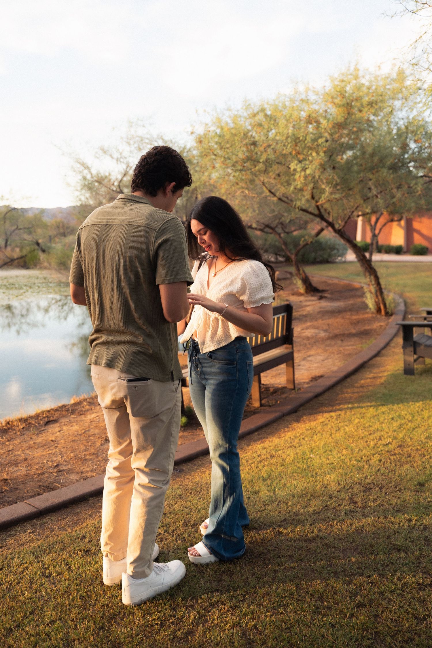 Two people walking together on a grassy path by a lake during sunset, viewed from behind.