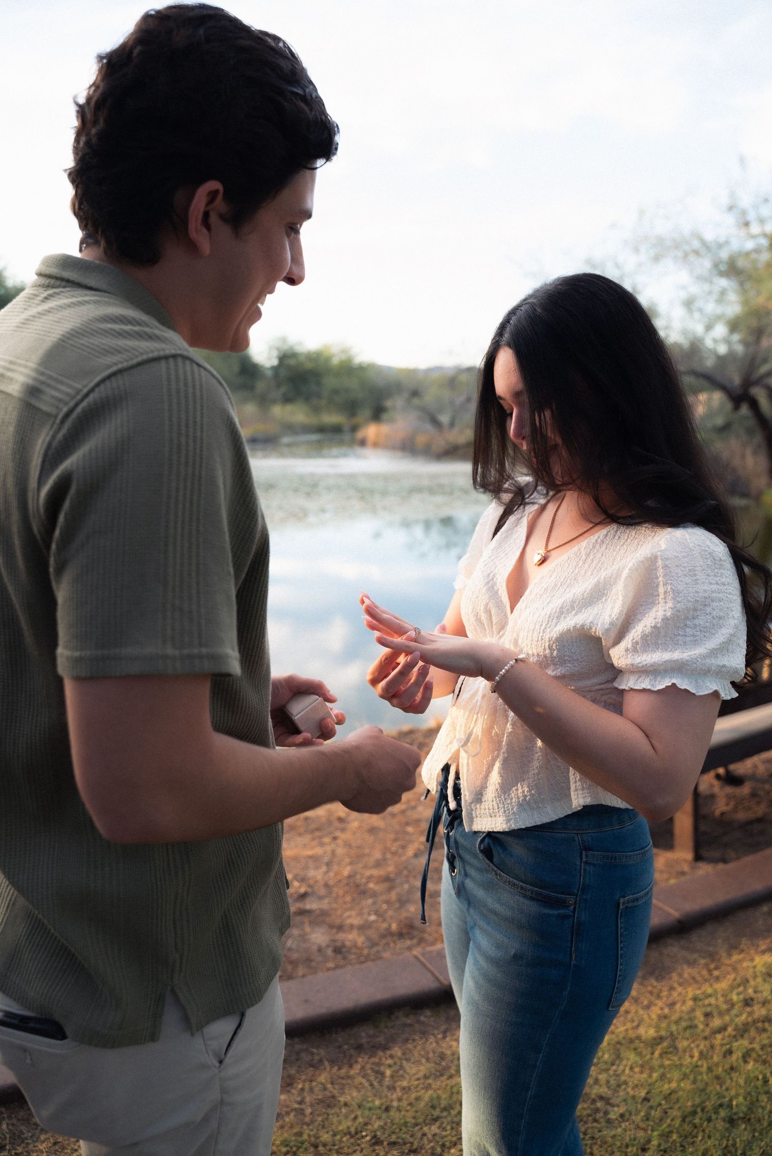 A couple stands together by a lake during sunset while looking at their phones.