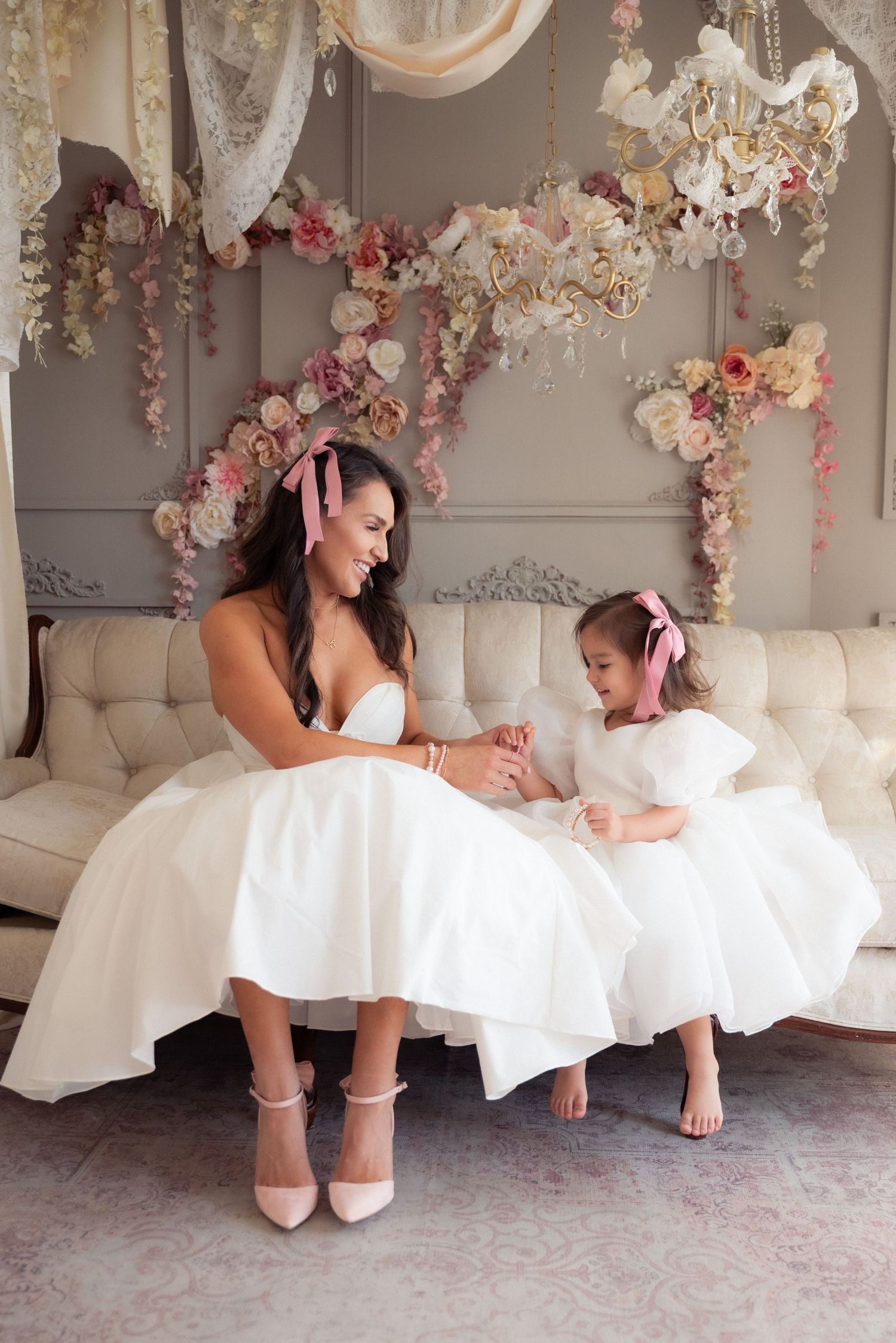 Two people in white dresses sit together on an elegant couch under floral decorations and a crystal chandelier.