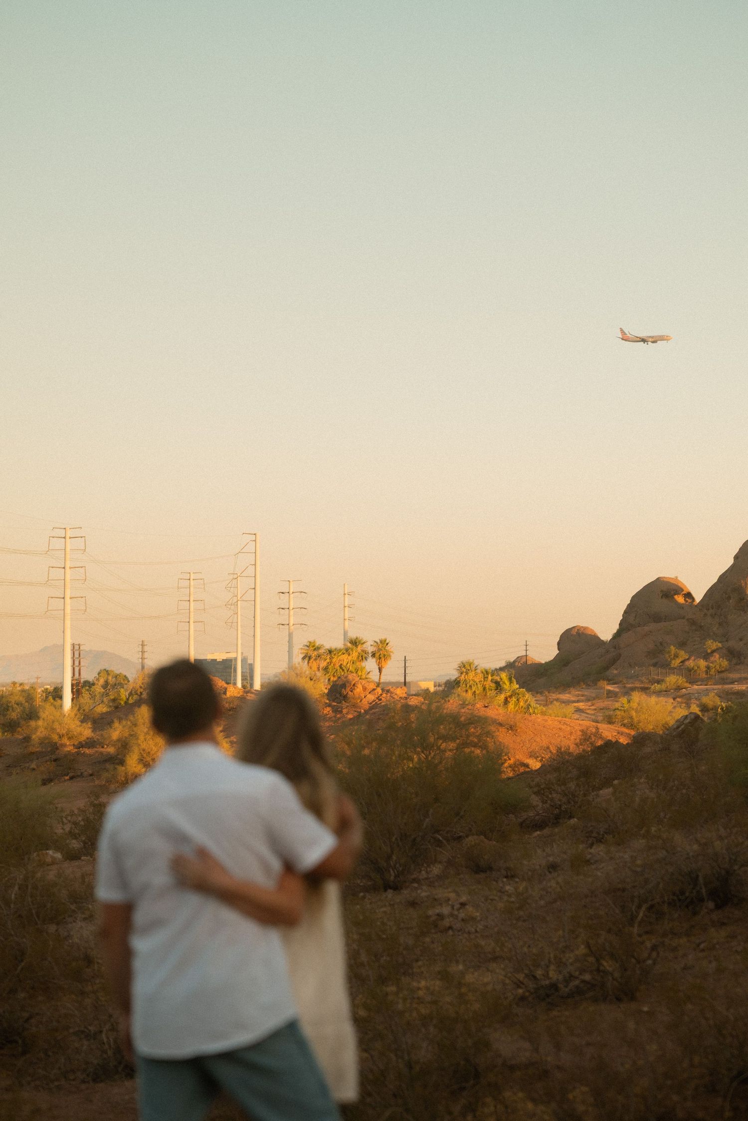 Couple embracing at golden hour in Arizona desert landscape with rocky formations and airplane in sky.