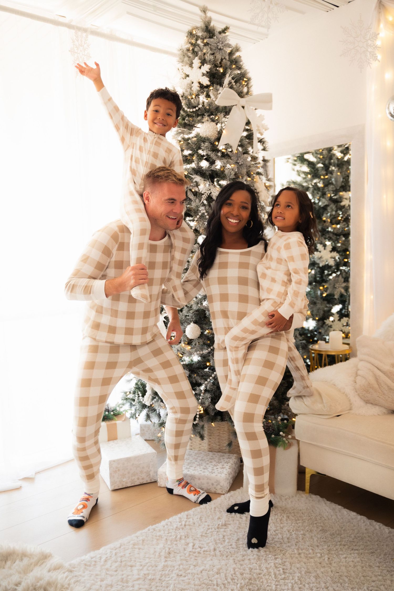 Family of four in matching plaid Christmas pajamas posing joyfully in front of a decorated holiday tree.