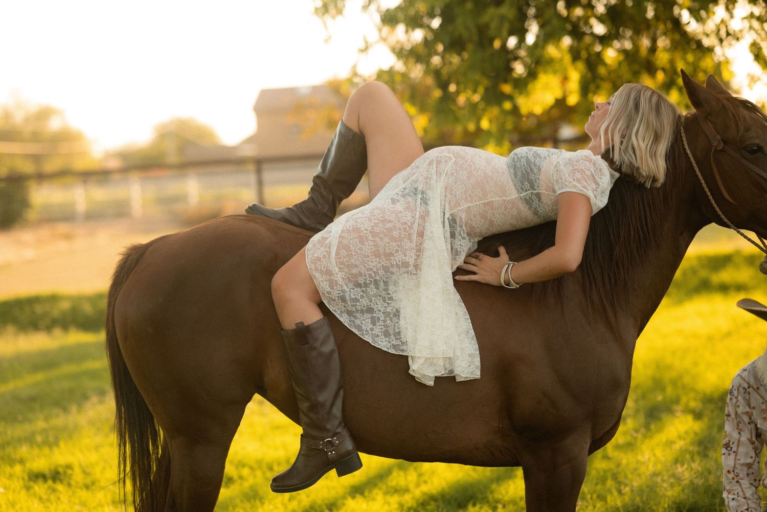 Woman in white lace dress and boots lying across a dark horse in golden sunset light on a green farm.