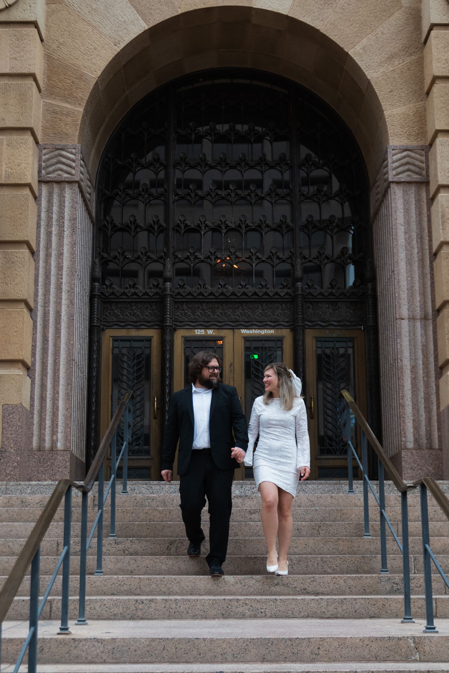 People walking up and down stone steps at the entrance of a historic building with arched doorways and handrails.