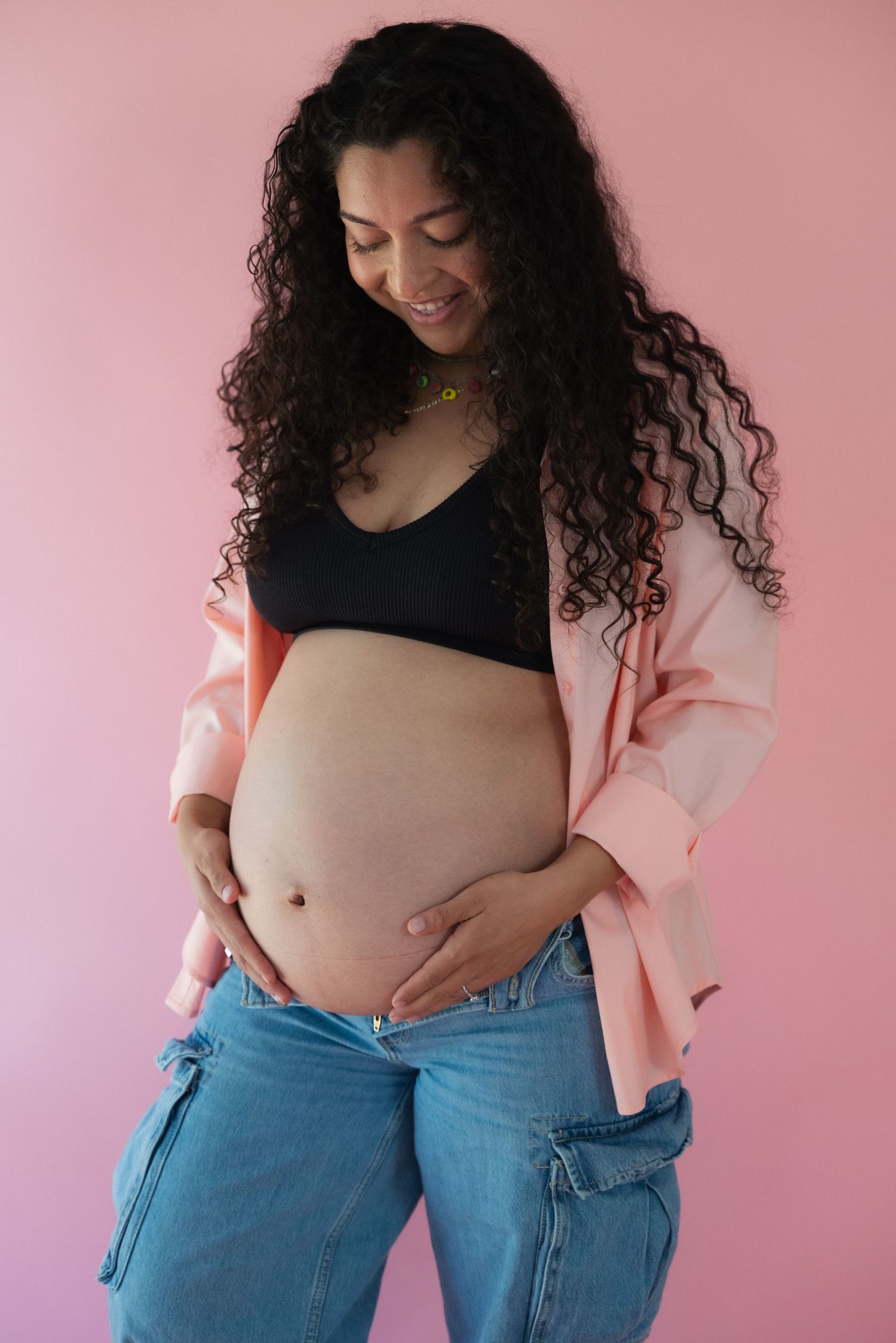 Maternity photo series showing pregnant woman in black top and jeans against pink background.