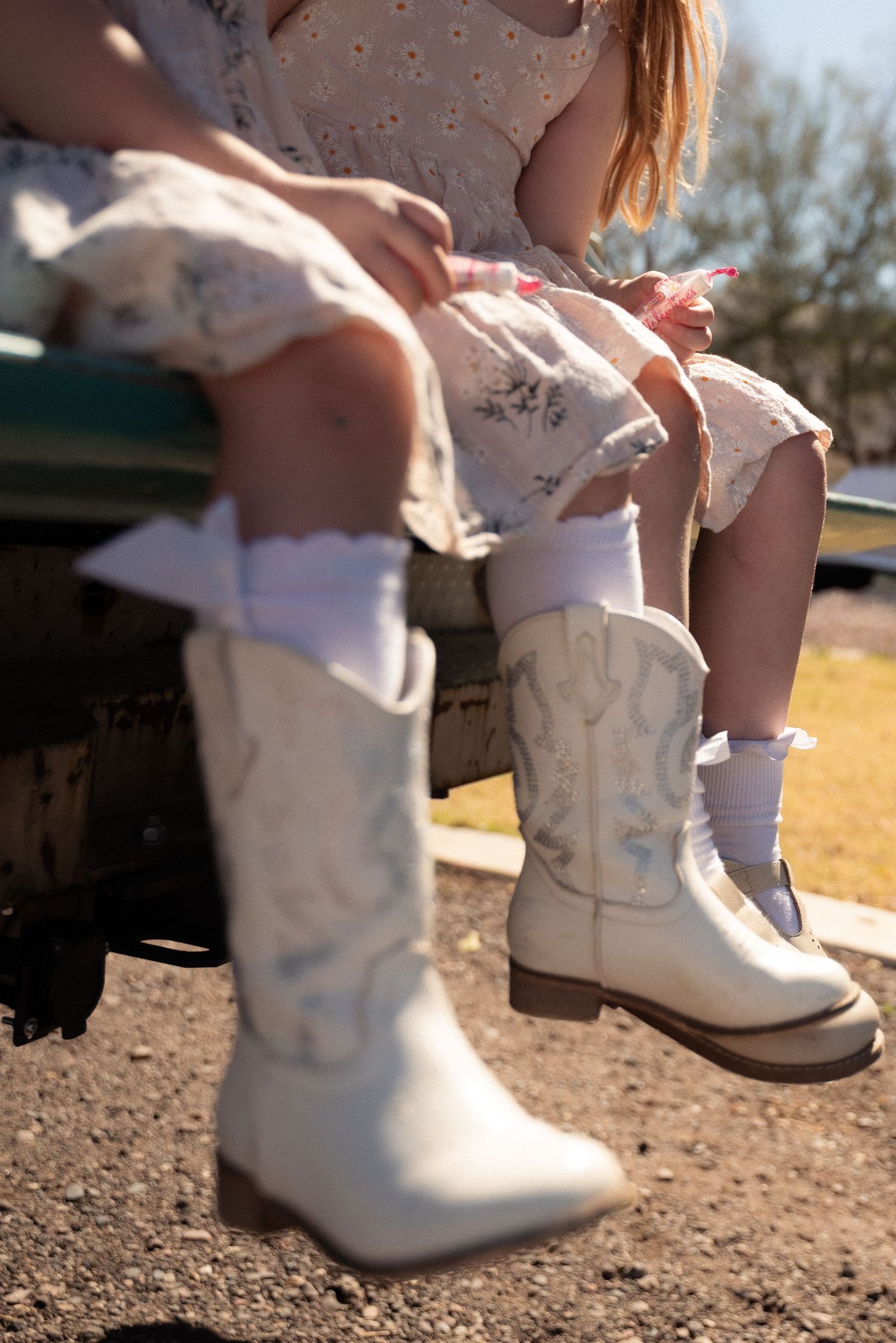 White cowboy boots and frilly dress detail shown in sunlit country setting with vintage truck in background.