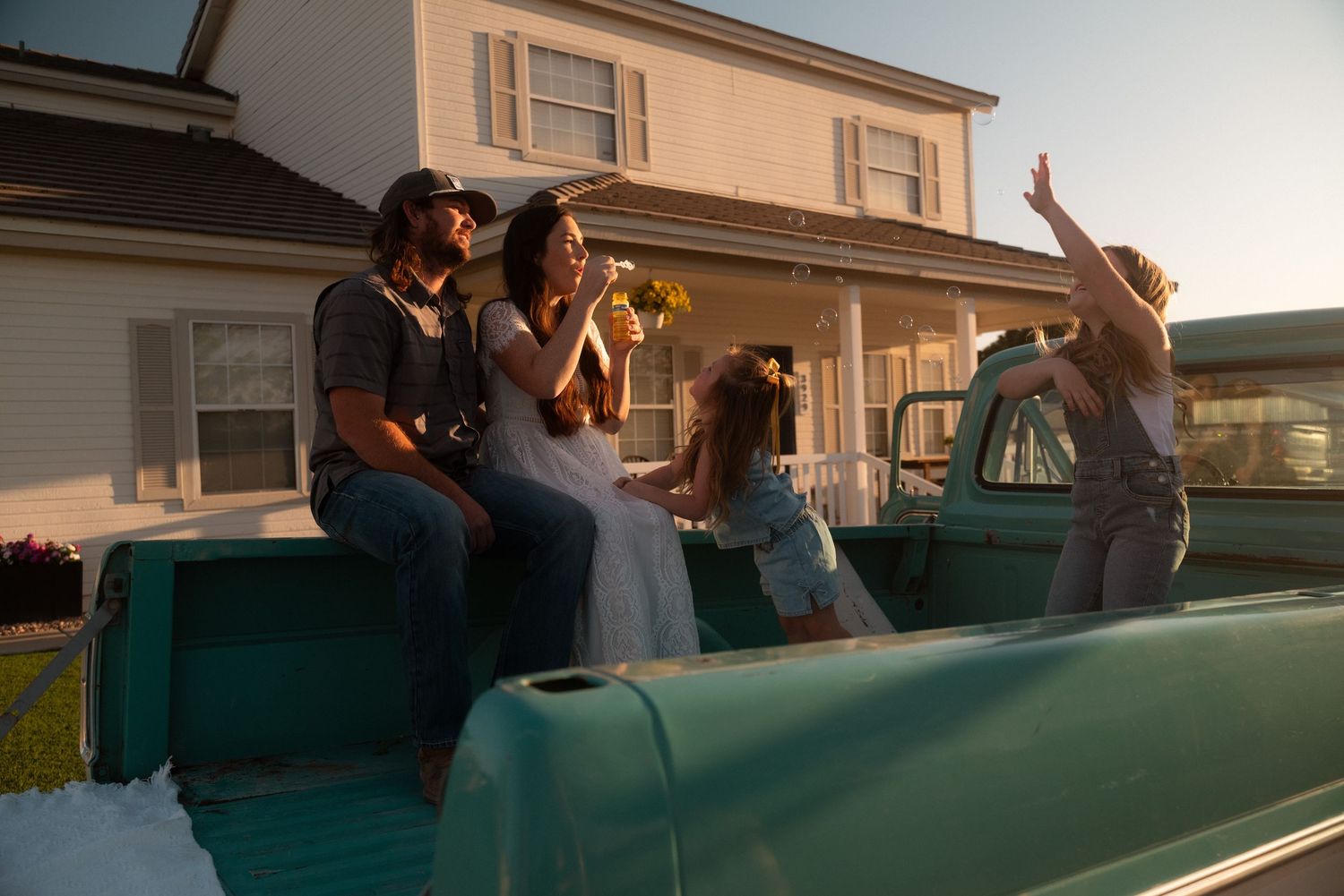 Wedding couple sitting on vintage teal truck bed with guests celebrating at golden hour outside farmhouse.
