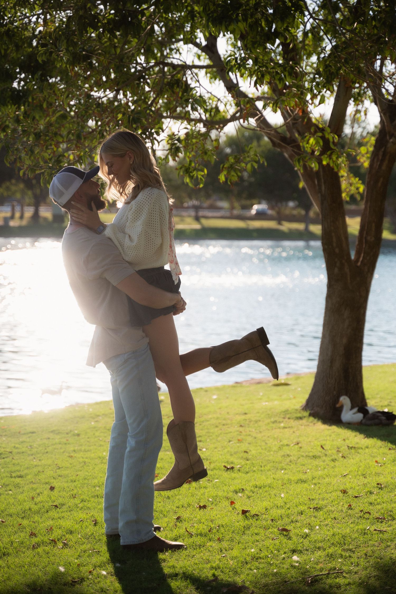 A romantic couple embraces under a tree by a serene lake during golden hour, creating a tender silhouetted moment.