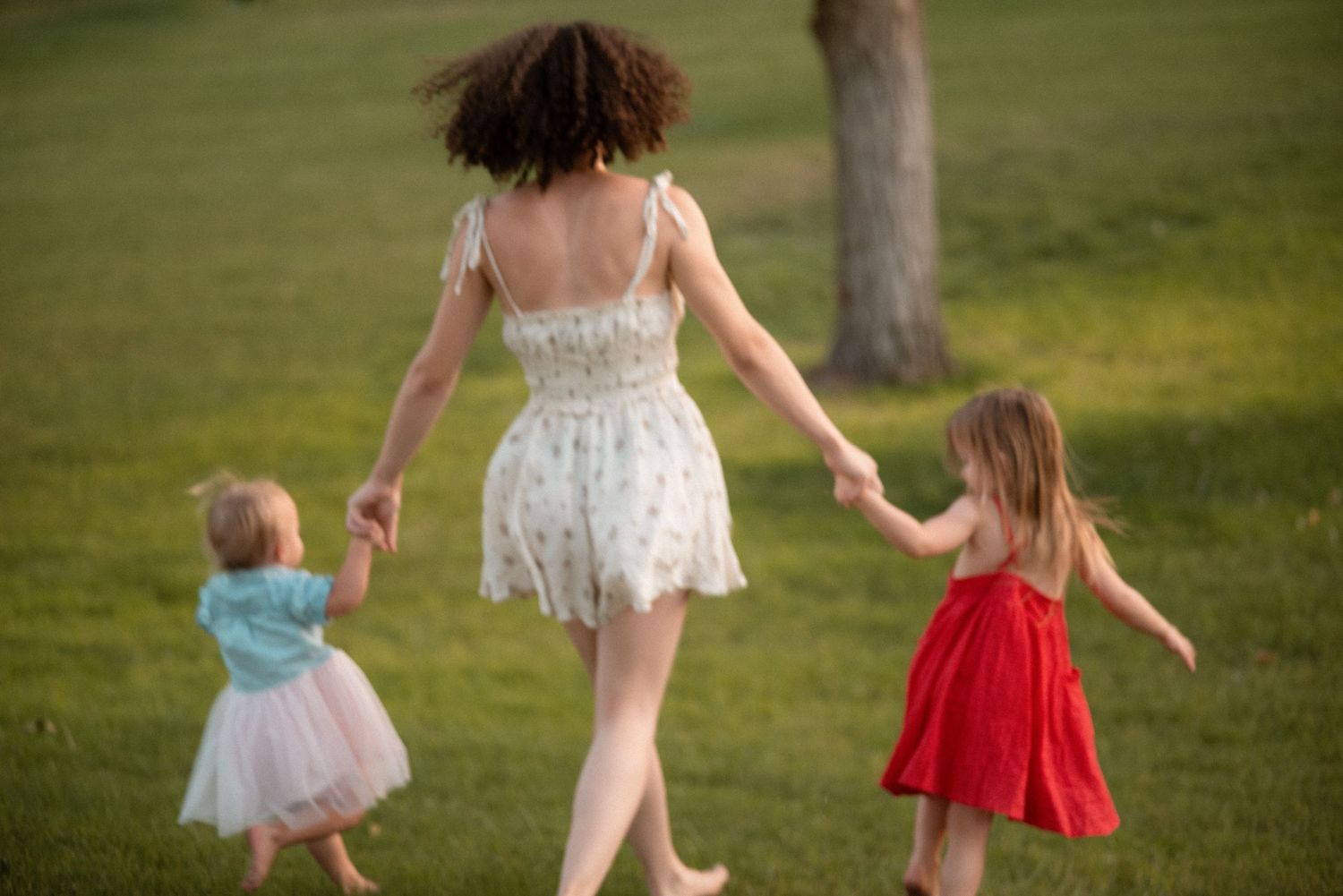 A woman in a floral dress holds hands with two little girls in colorful dresses while walking barefoot on green grass.