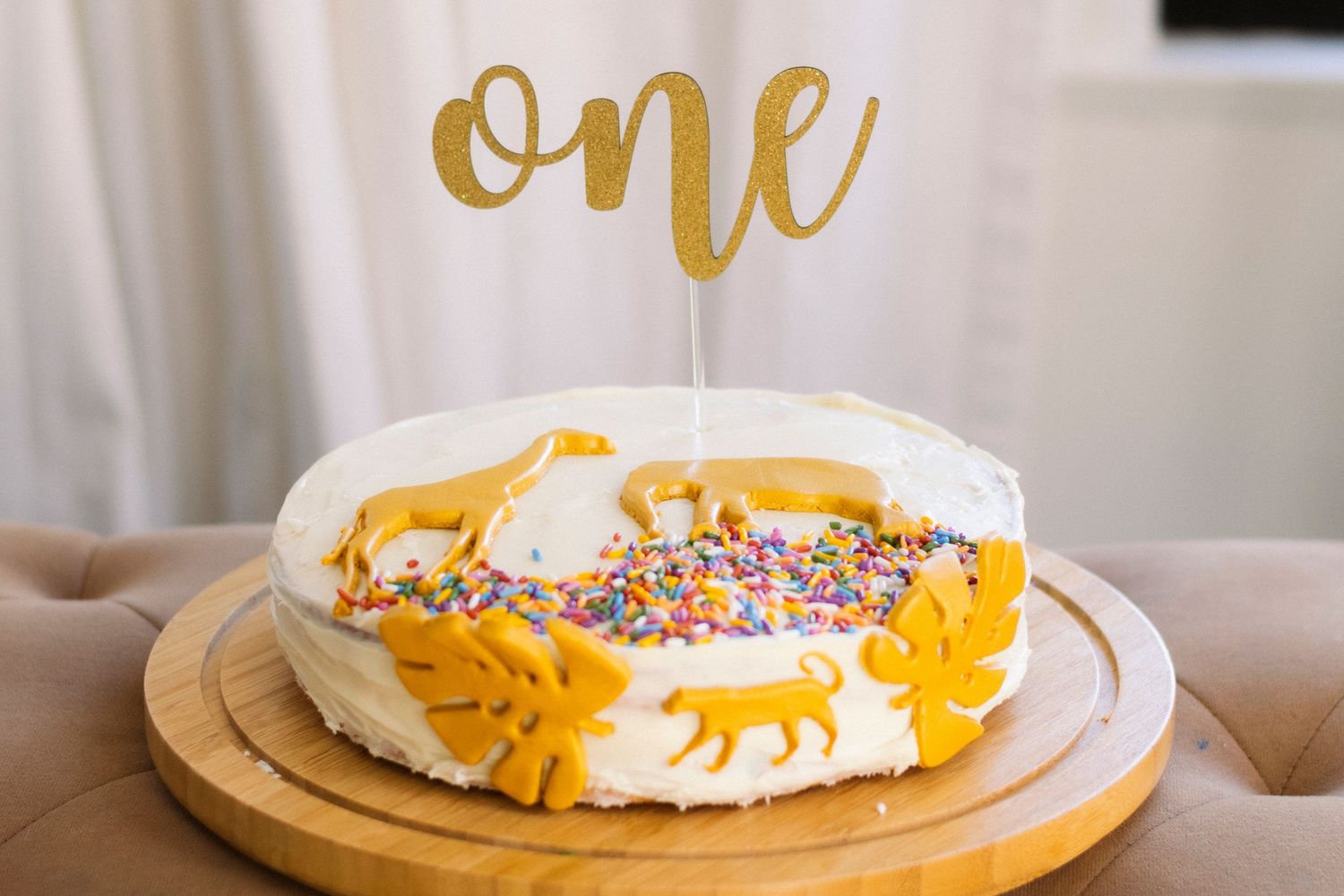White frosted birthday cake with yellow floral decorations and gold 'one' topper sits on wooden serving board.