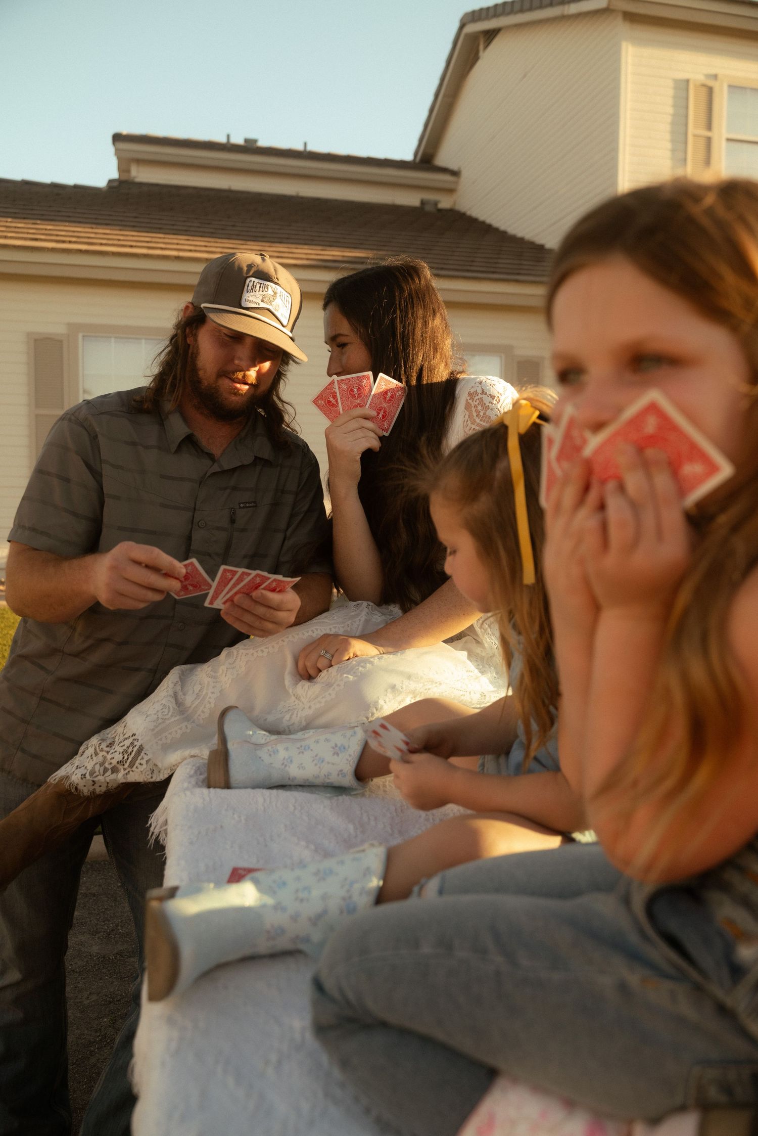 Family playing cards together outdoors near a house during golden hour sunset.