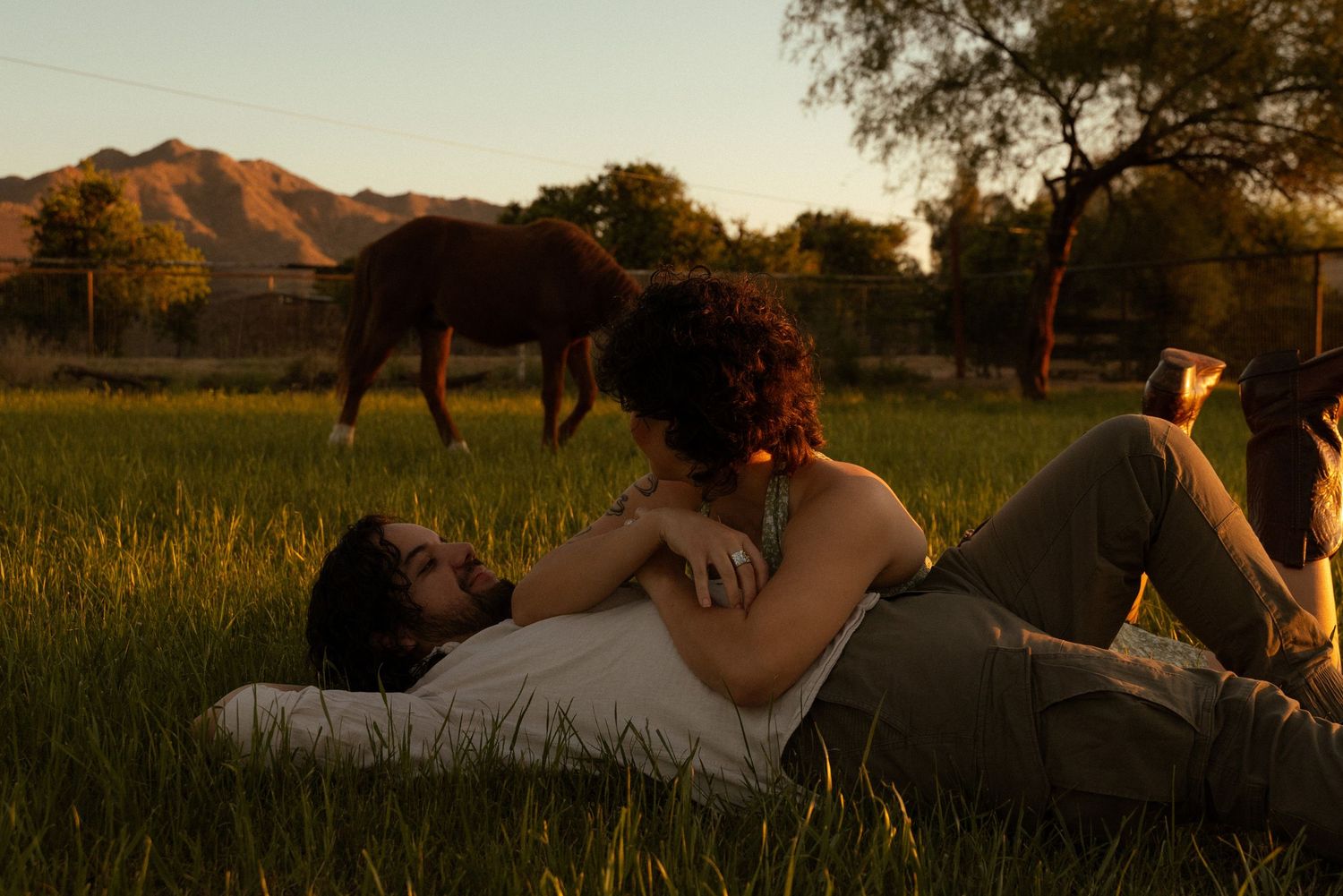 Couple lying in green grass at golden hour with horse grazing and mountains in background.