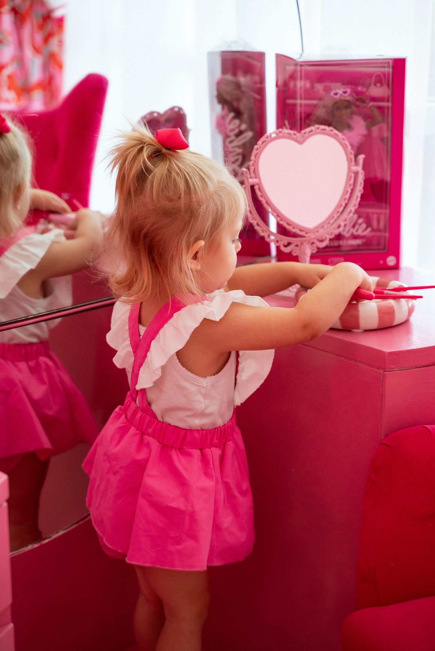 A small child in a pink dress stands at a vanity table with a heart-shaped mirror and pink decorative elements.