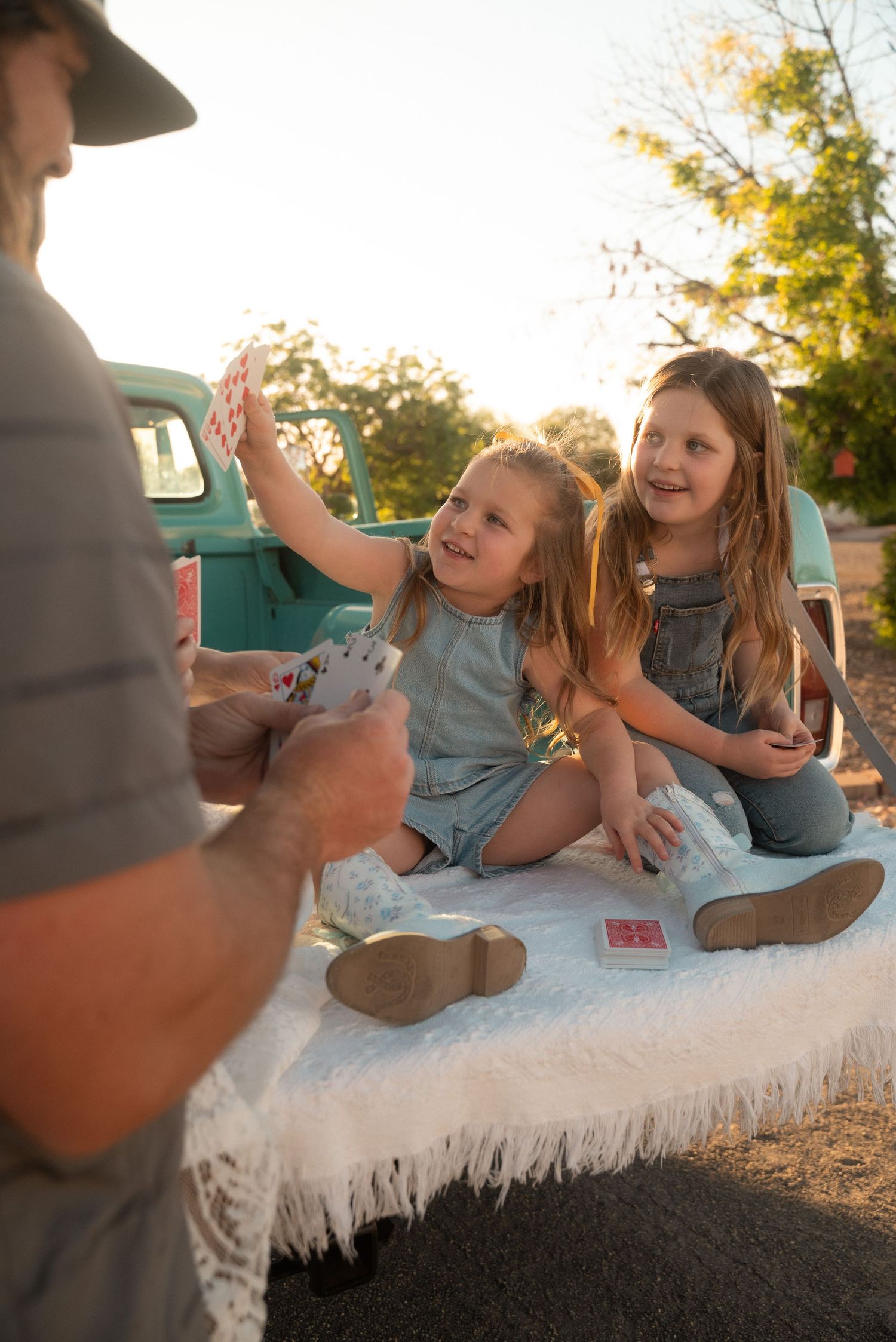 Two young girls in denim outfits play cards with an adult on a blanket near a vintage teal truck outdoors.