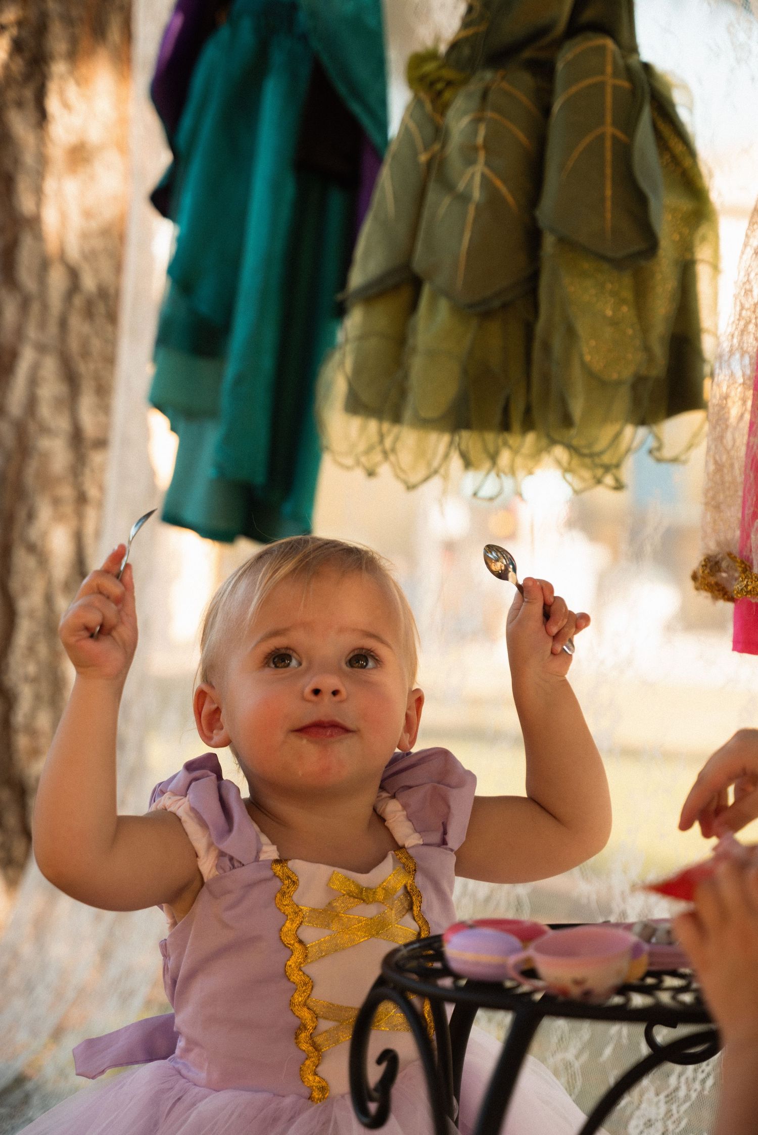 Toddler girl in purple princess dress holds spoons up at outdoor tea party with fairy costumes hanging behind her.