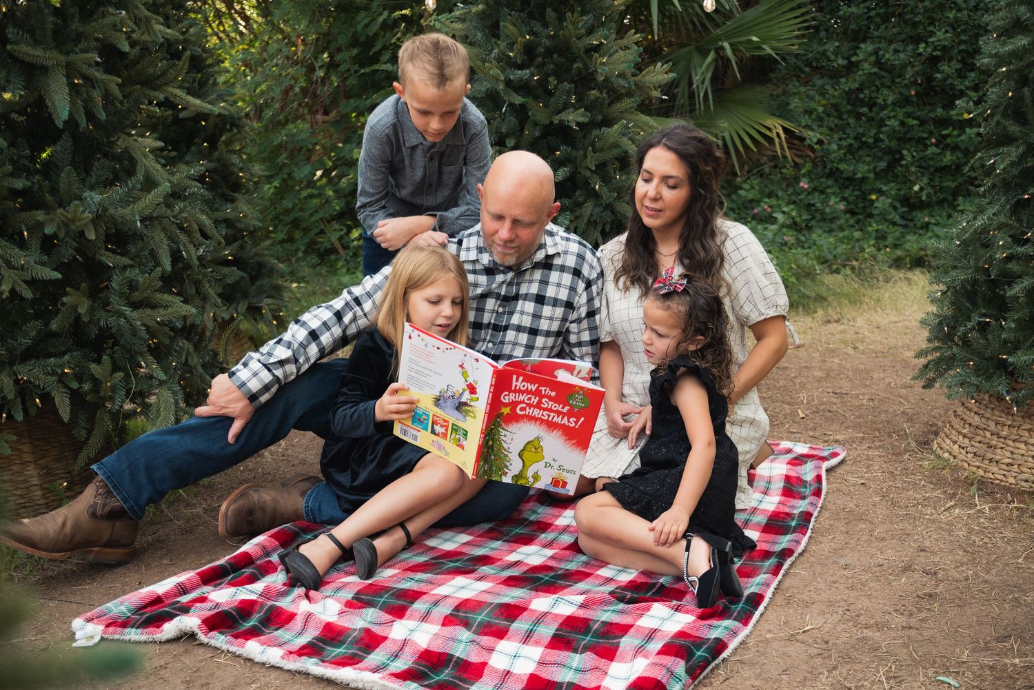 Family gathered on plaid blanket among Christmas trees reading a holiday book together outdoors.