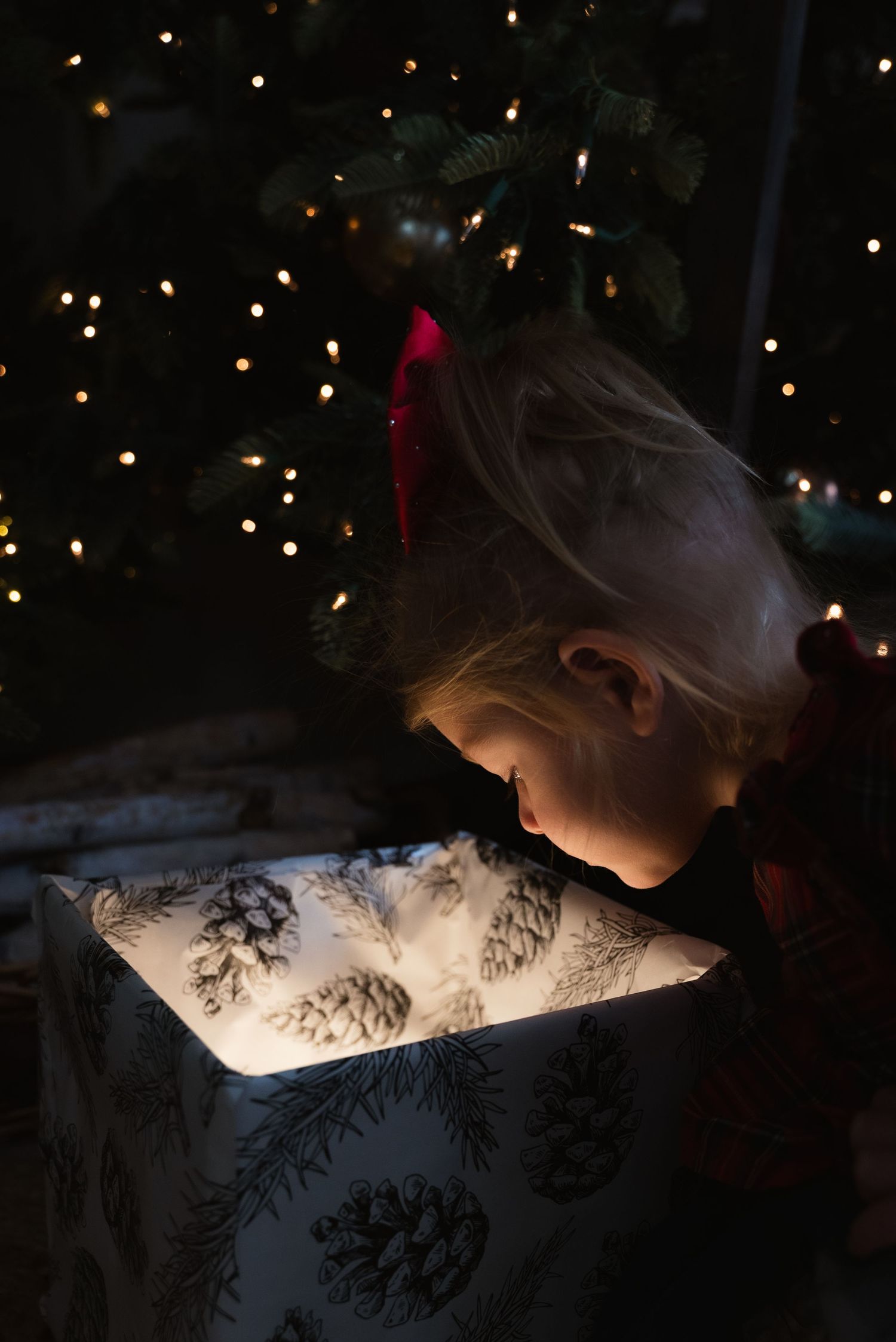 A young child with a red bow peers into a glowing gift box near a lit Christmas tree in a dark room.