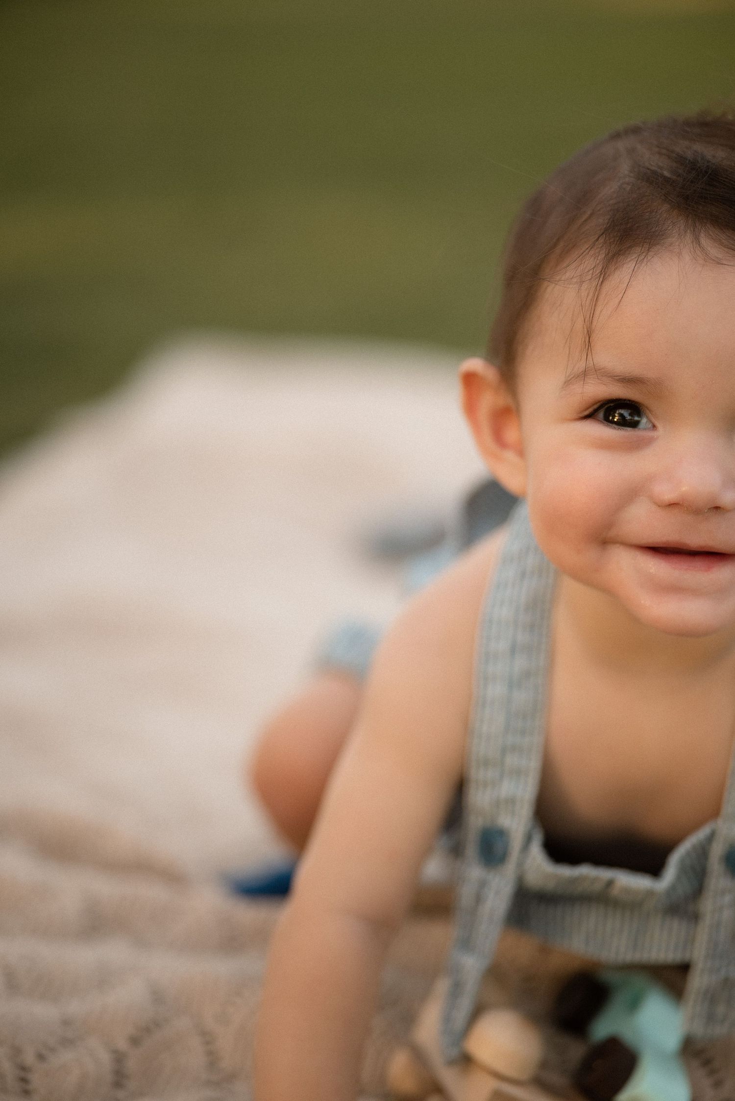 Smiling baby boy in striped overalls lying on a blanket outdoors on green grass.
