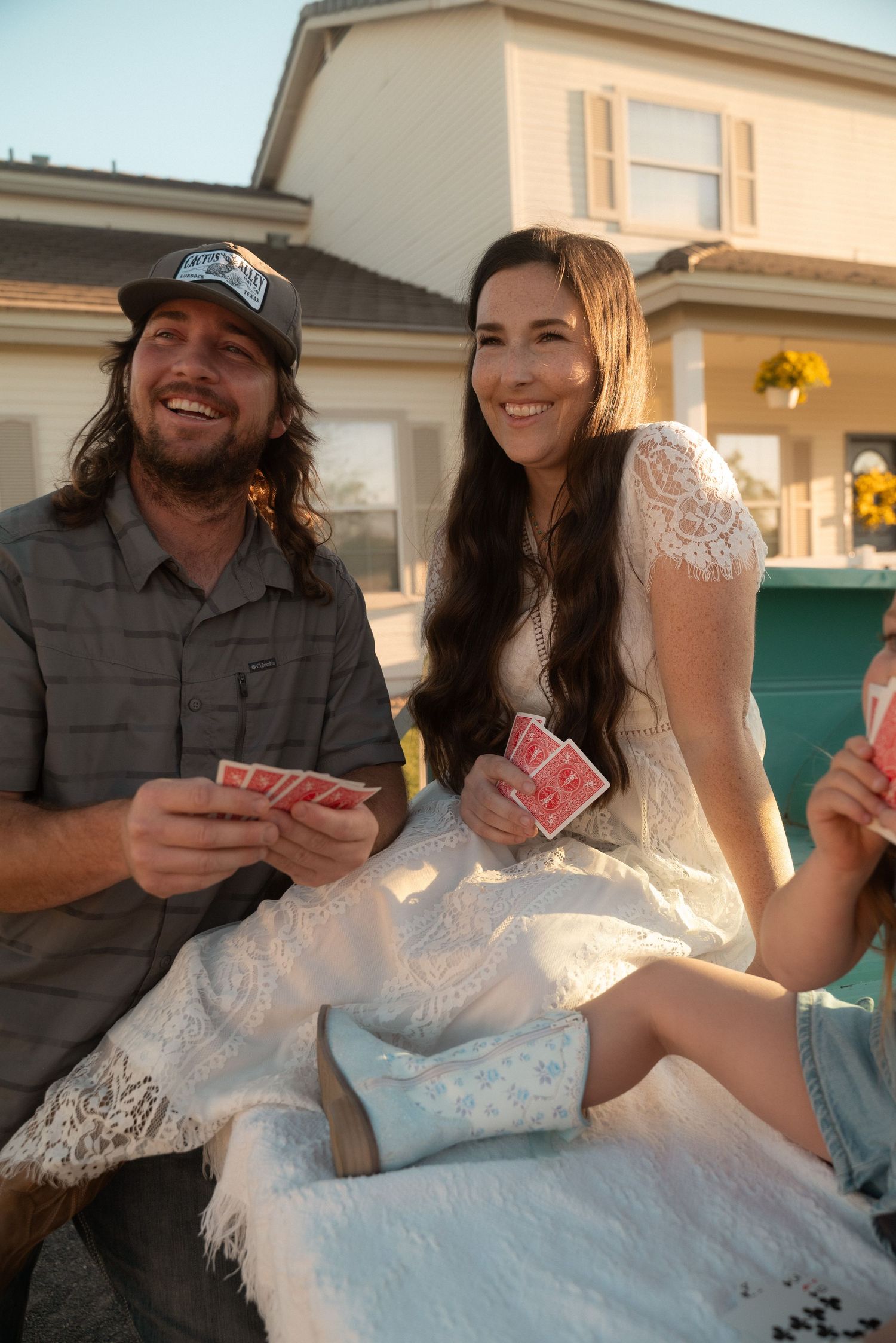 Smiling bride in white lace dress and groom in gray shirt playing cards outdoors at sunset wedding reception.