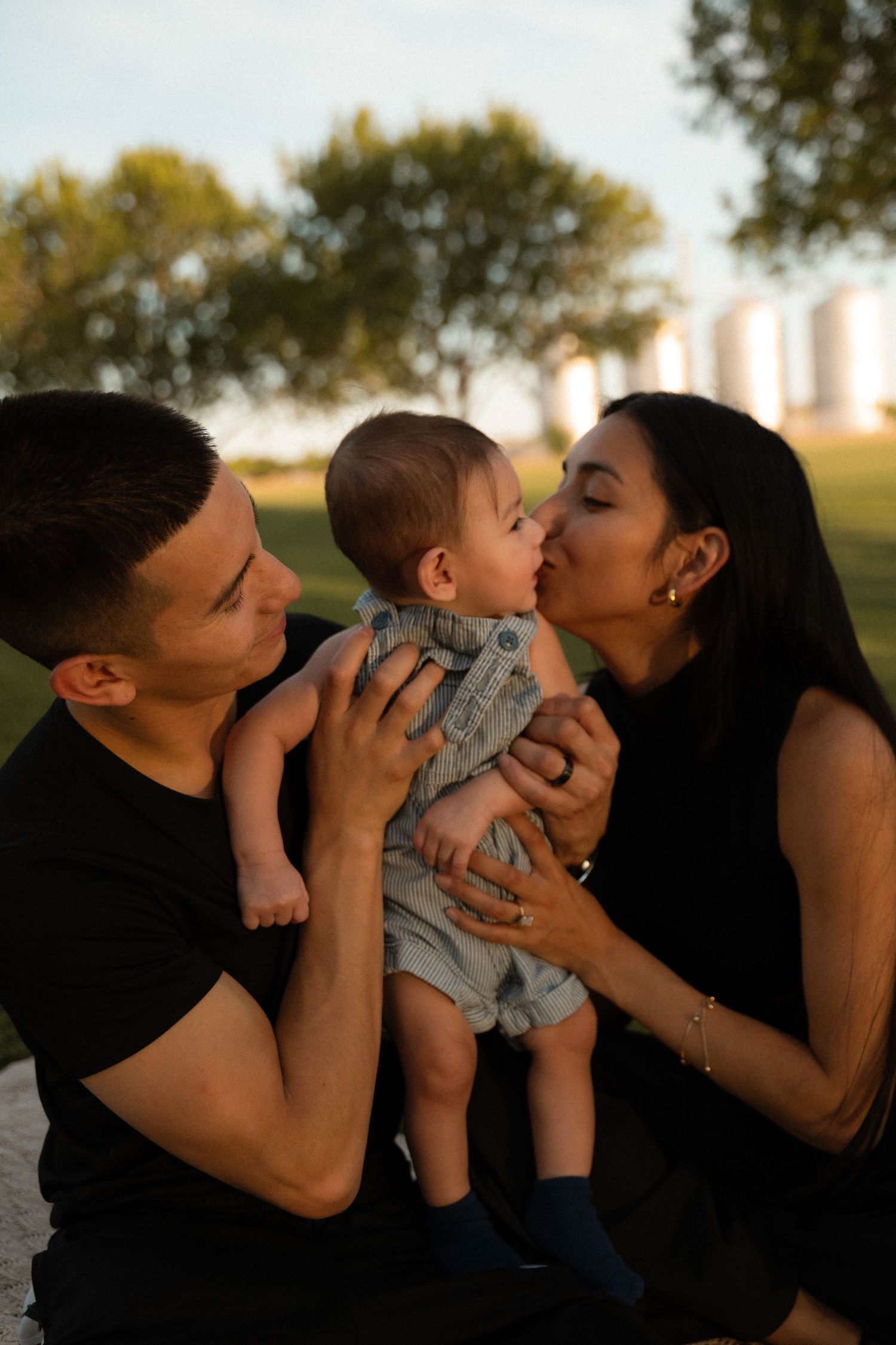 A young family shares a tender moment outdoors at golden hour, with parents kissing their baby boy in a park.