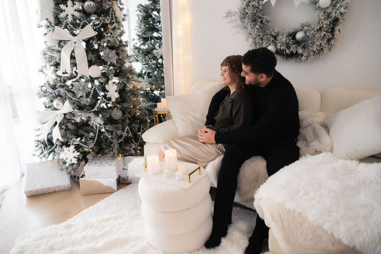 A couple sits together in a cozy white room decorated for Christmas with a flocked tree and silver wreath.