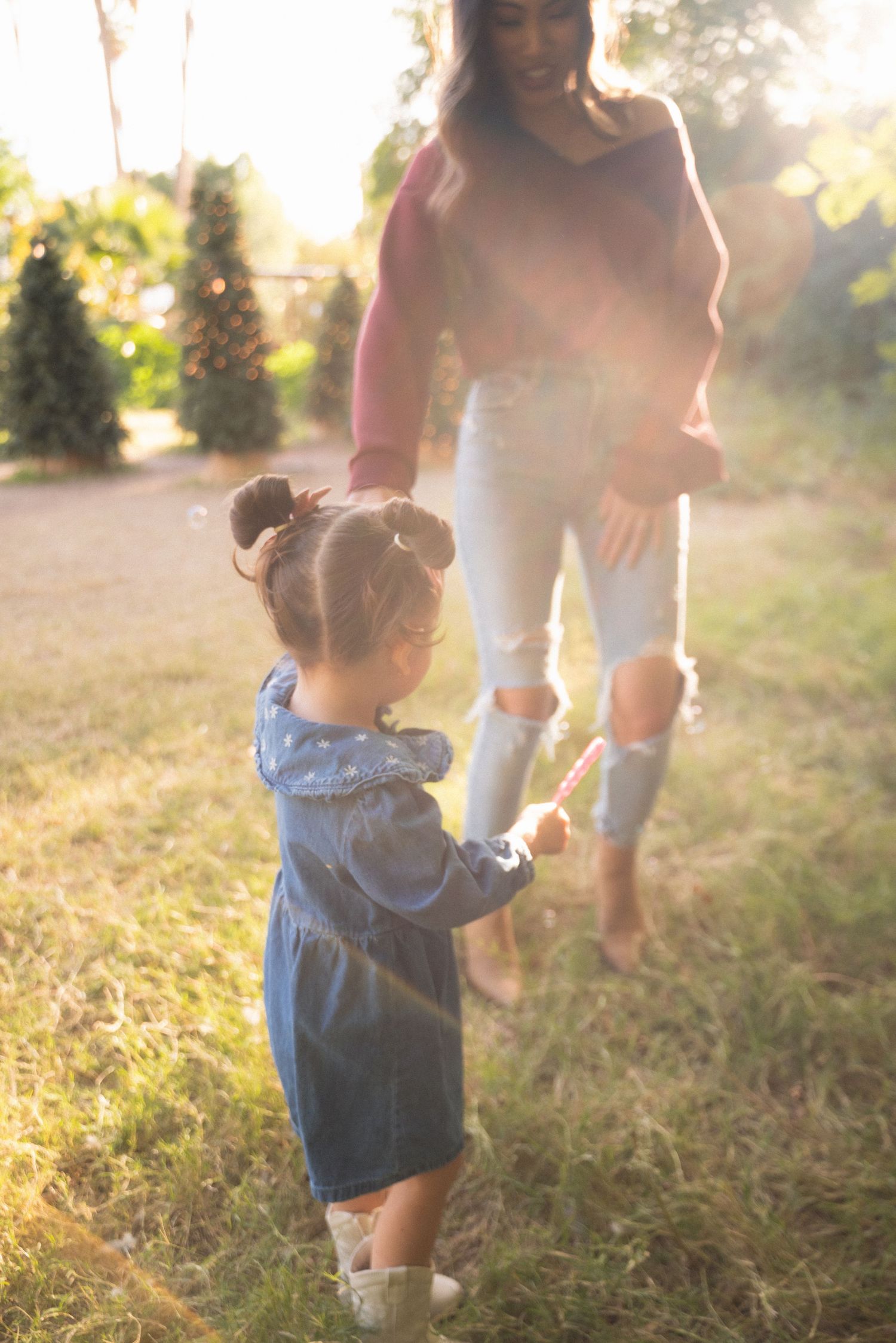 A young girl in a denim dress holds hands with a woman in a sunlit outdoor setting.