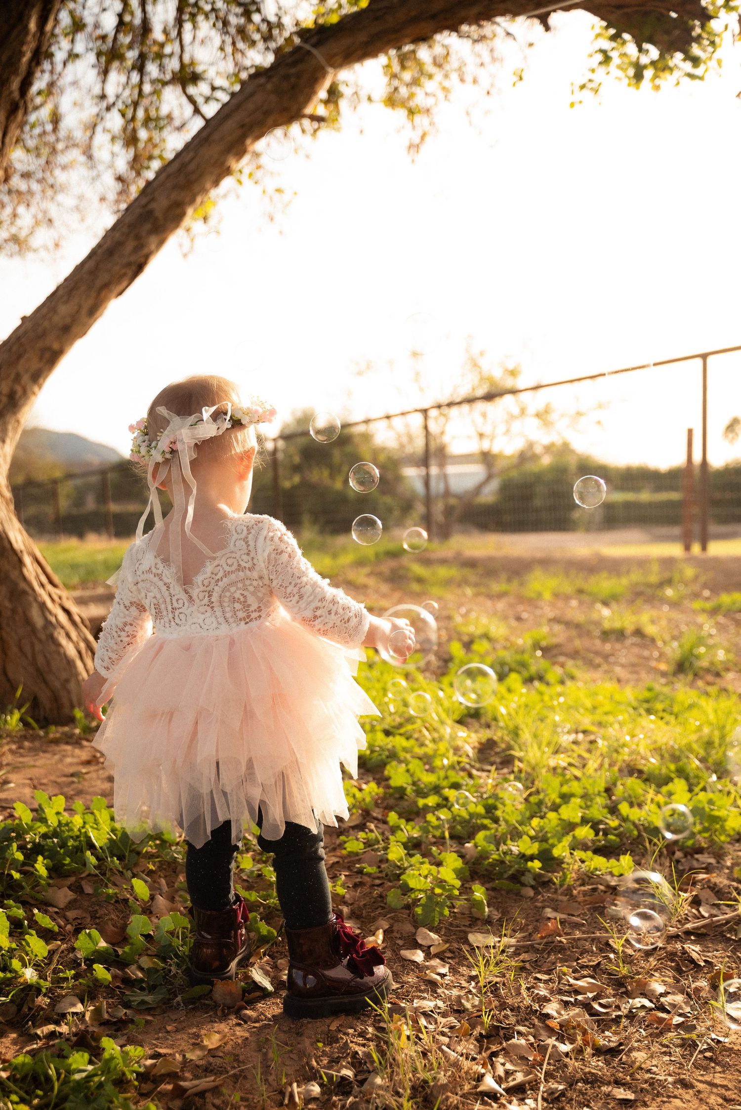 A small figure in a pink tulle dress and boots plays with bubbles in a sunlit garden at golden hour.