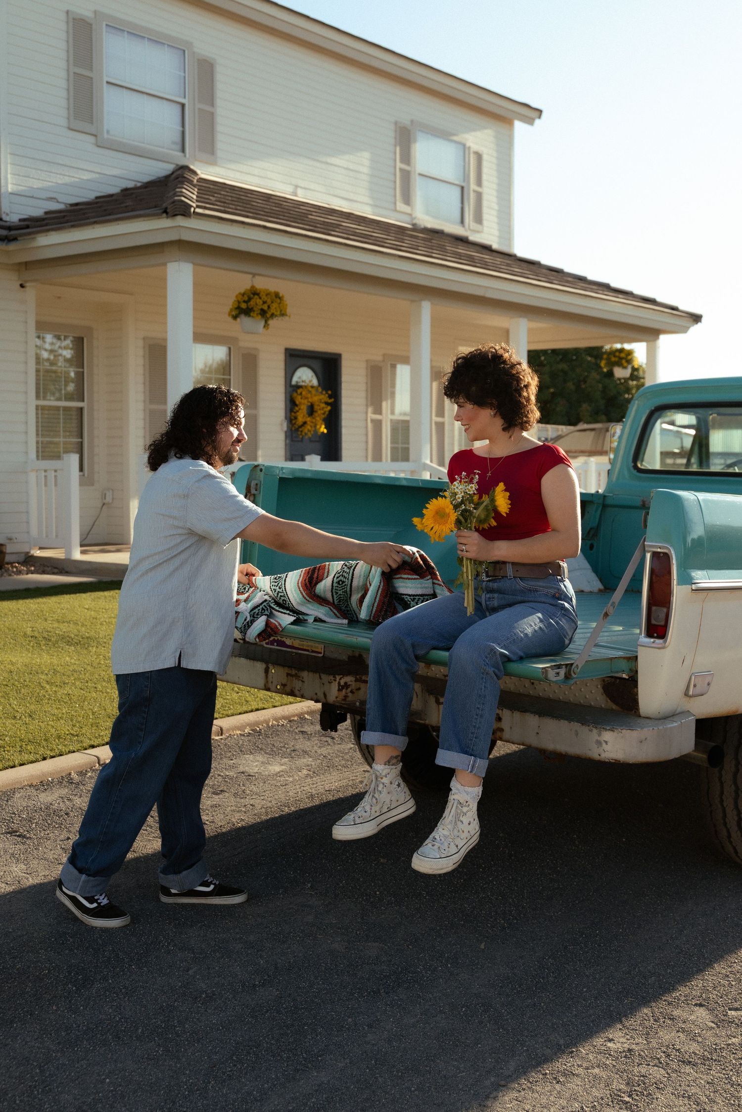 Two people exchanging sunflowers by a vintage teal pickup truck outside a white farmhouse.