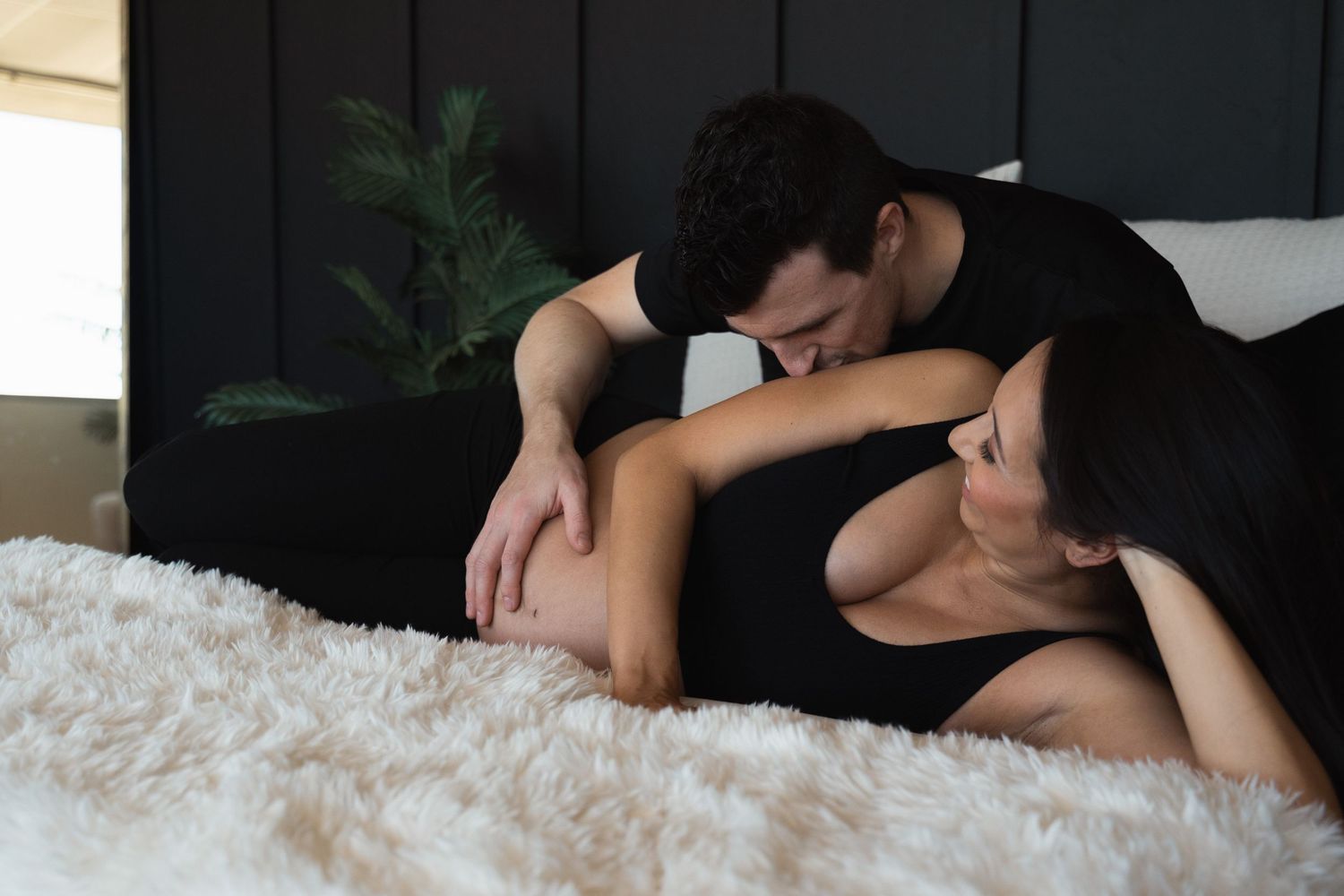 A couple shares a tender moment on a bed, with a man kissing a pregnant woman's belly.