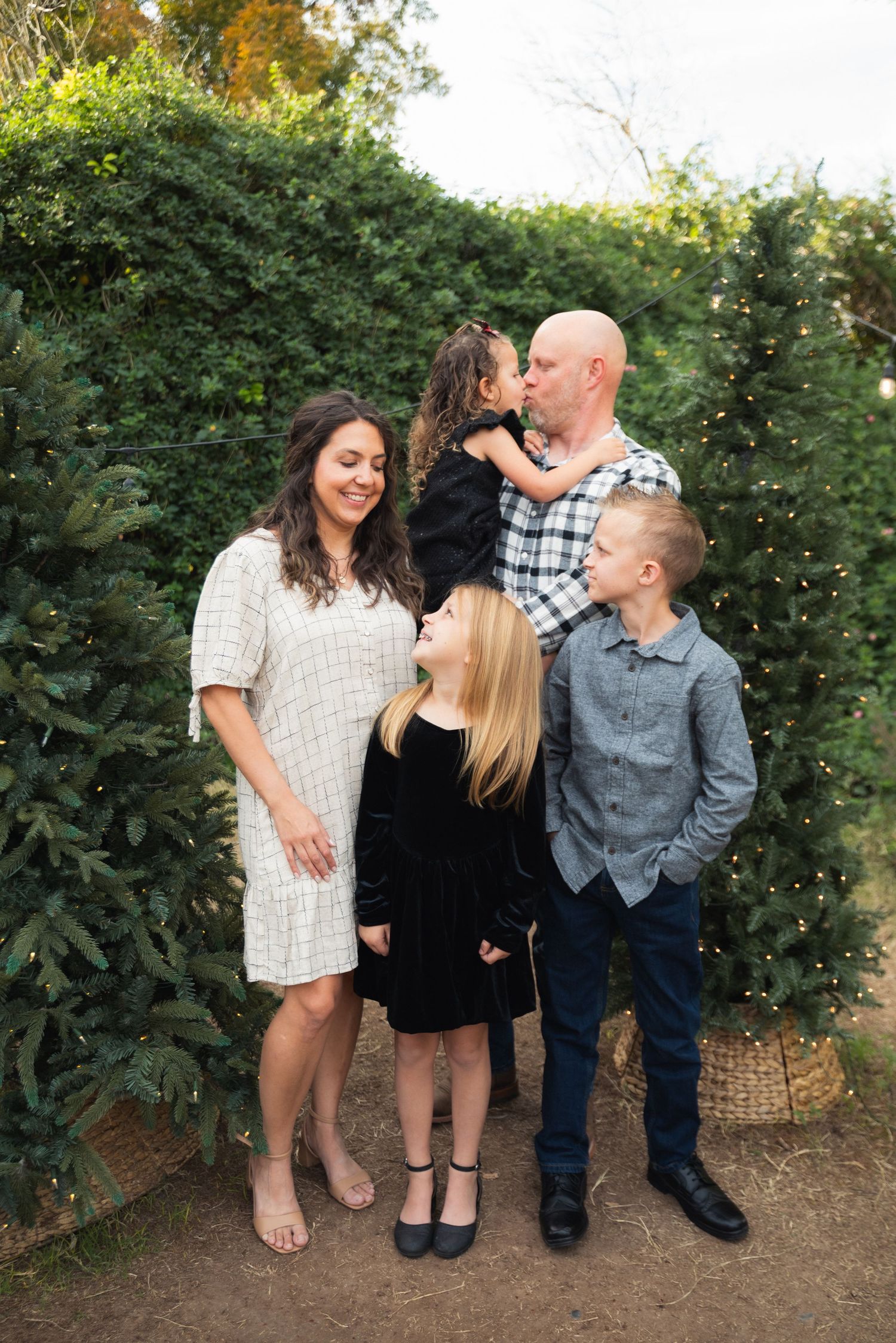 Family of five posing outdoors among lush green hedges, parents with three children dressed in holiday attire.