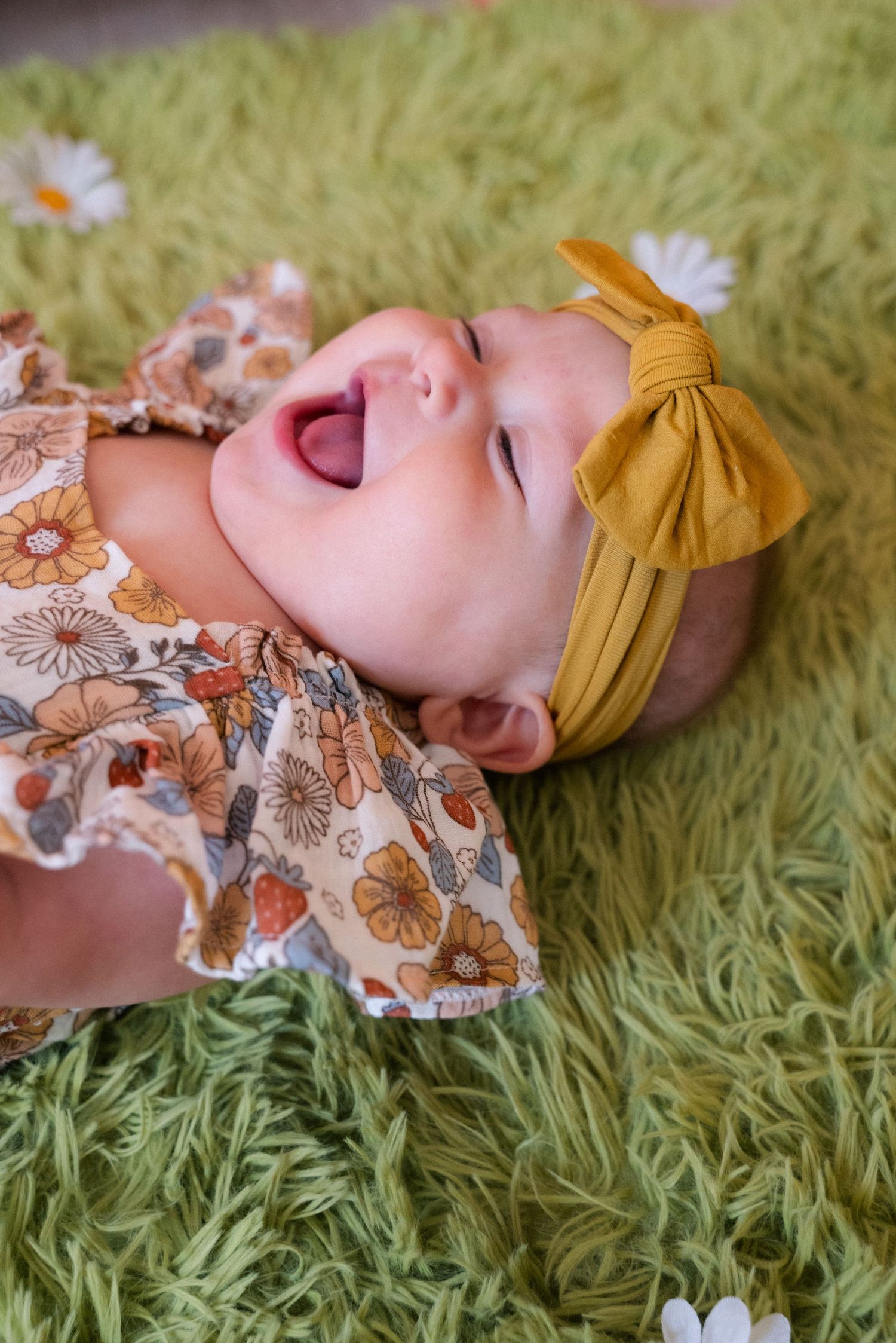 A baby lying on green shag carpet wearing a mustard yellow headband and floral dress while looking up with joyful expression.