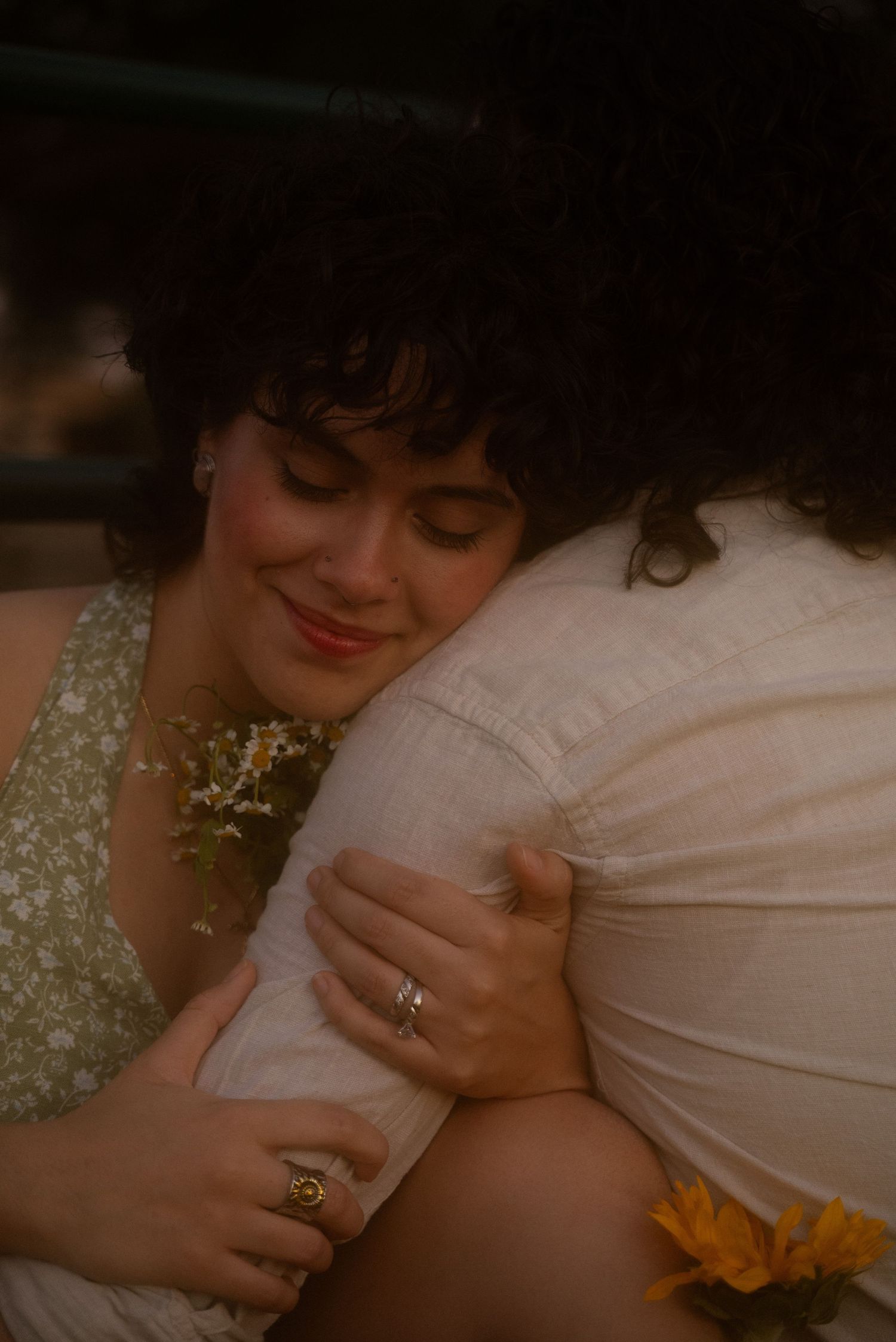 Two women with curly dark hair embrace tenderly, one wearing a floral dress and rings, holding small white flowers.