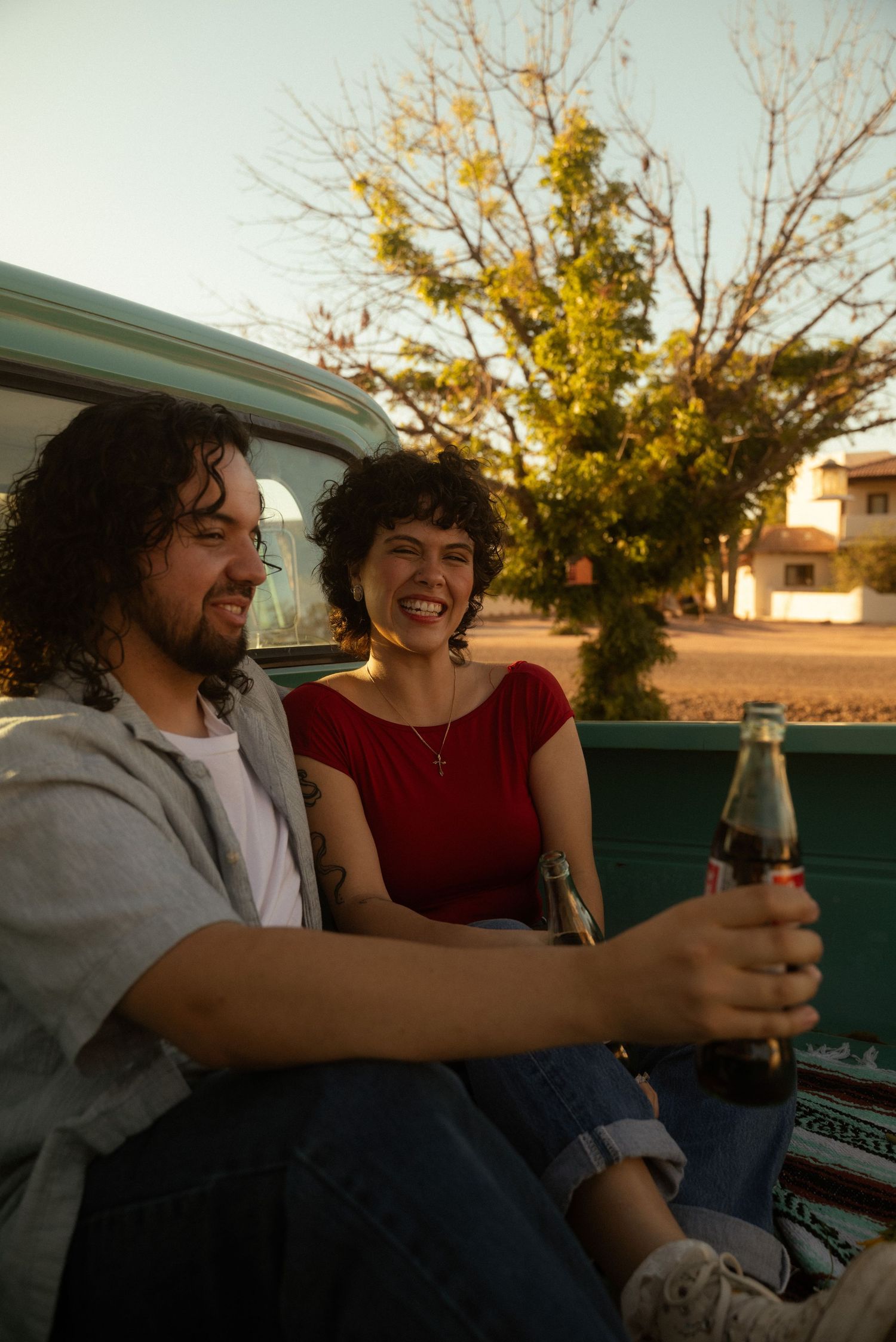 Two young adults sitting in a pickup truck bed, smiling and holding soda bottles at sunset.