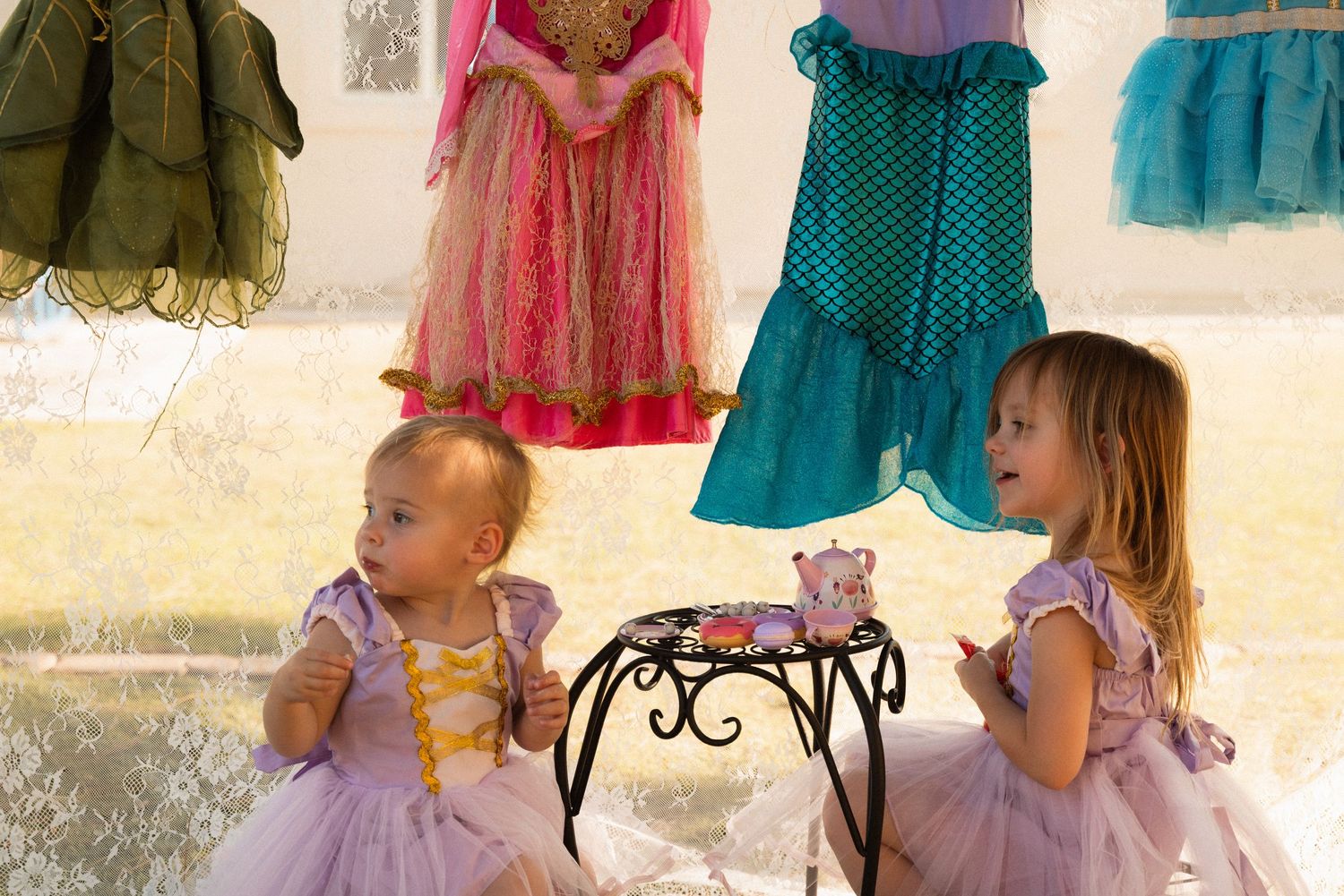 Two young girls in purple princess dresses enjoy a tea party outdoors with colorful costumes hanging in the background.