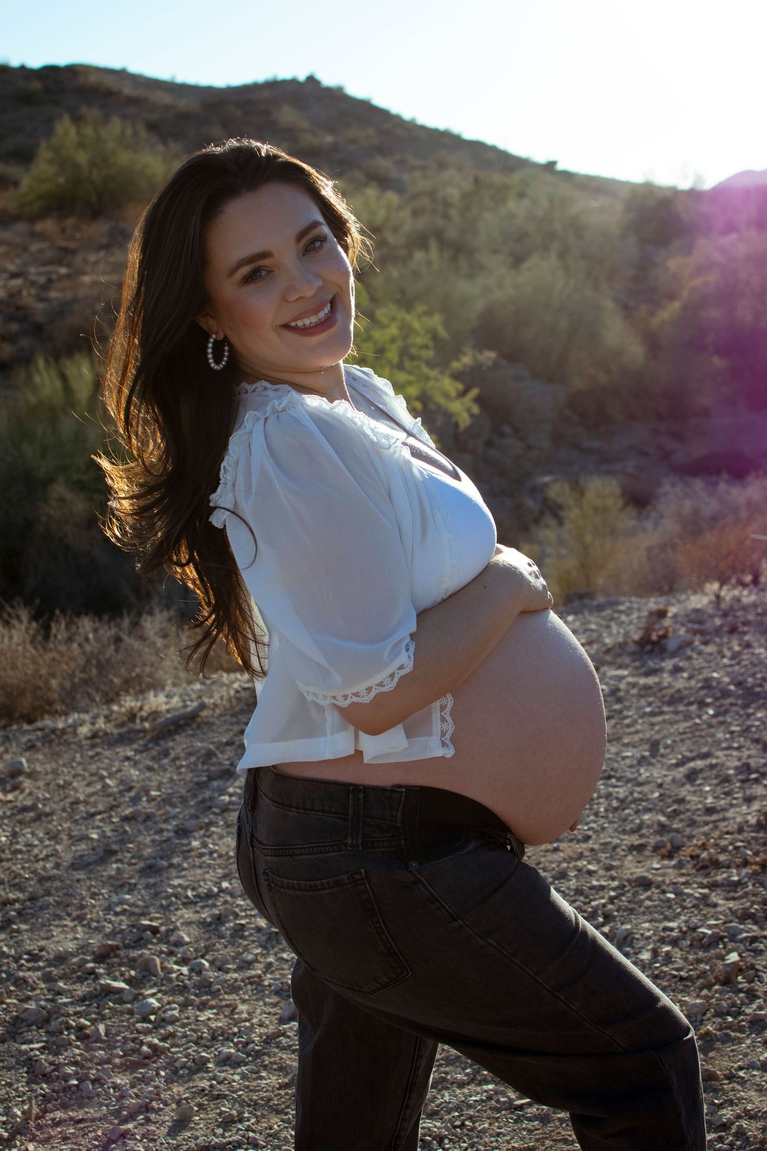 A pregnant woman in a white top poses against a desert mountain backdrop during golden hour.